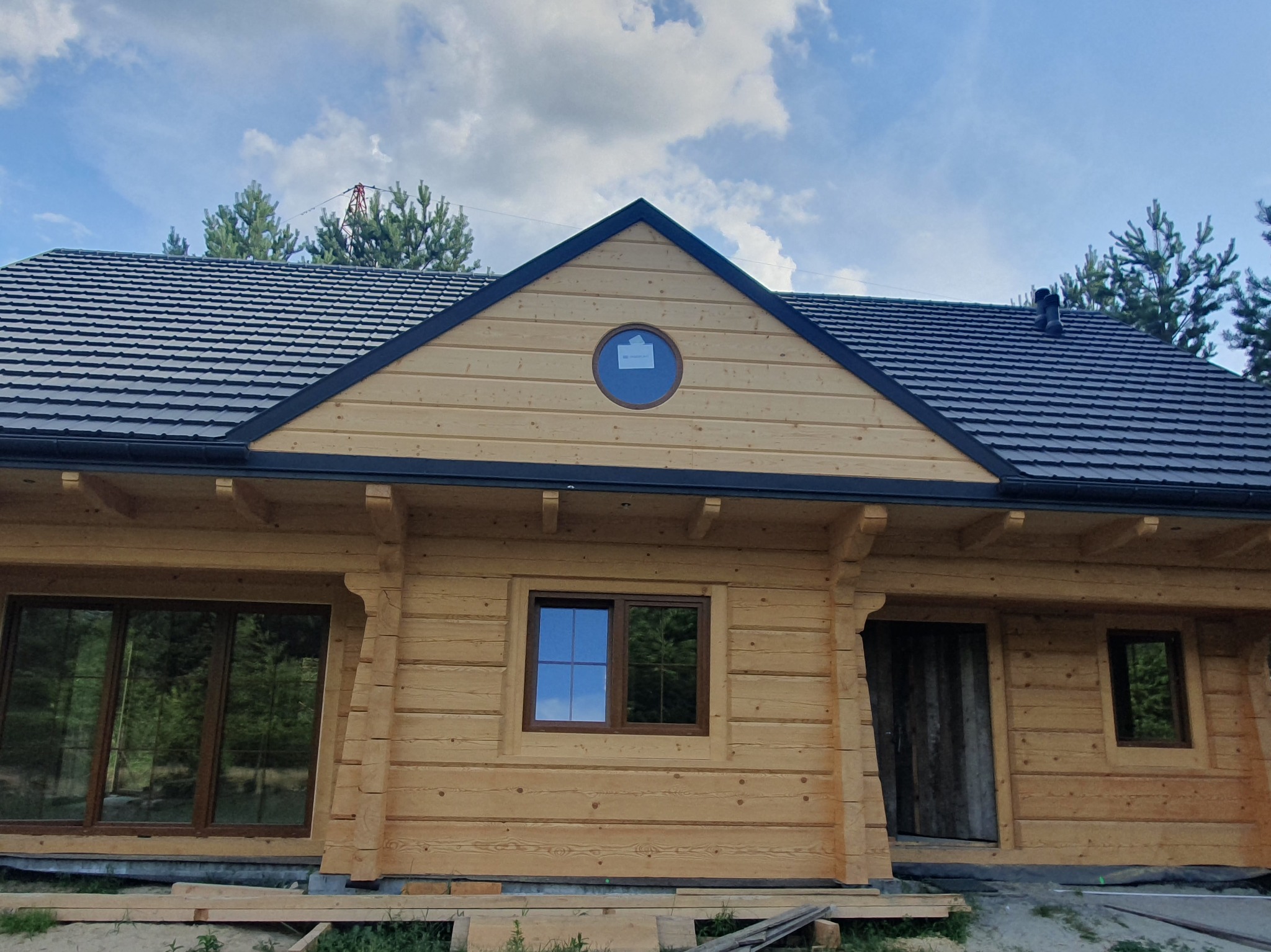 Surowy dom z bali z ciemną dachówką i okrągłym oknem na szczycie. Widoczne elementy konstrukcyjne i wykończeniowe. Częściowo wykończona elewacja.