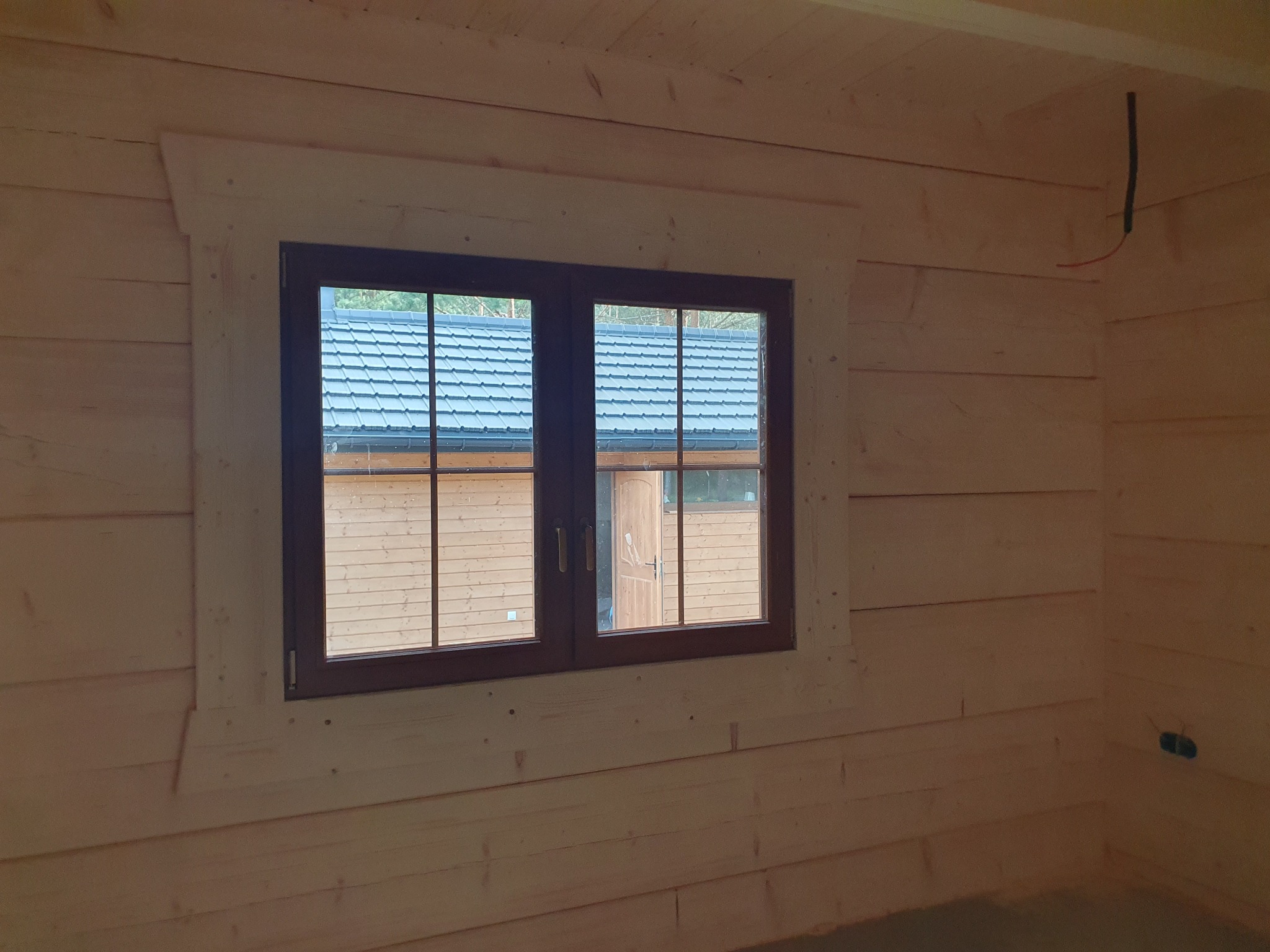 Wnętrze domu z bali w trakcie budowy: widok na okno z drewnianą ramą, przez które widać fragment dachu innego budynku.