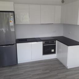 Mebleak-Meble na wymiar - Nowoczesna, minimalistyczna kuchnia z białymi, lakierowanymi frontami, czarnym blatem i szarą lodówką LG, wykończona panelami podłogowymi imitującymi drewno.