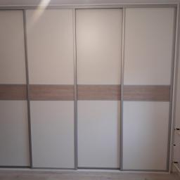 Mebleak-Meble na wymiar - Czterodrzwiowa szafa przesuwna z białymi frontami i poziomym pasem w kolorze jasnego drewna, widoczny grzejnik obok.