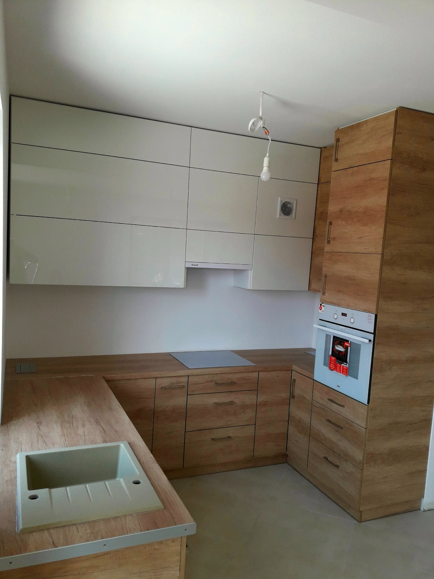 Jasna kuchnia z drewnianymi szafkami dolnymi i białymi górnymi, z widocznym zlewozmywakiem i piekarnikiem, niedokończone podłączenie oświetlenia sufitowego.