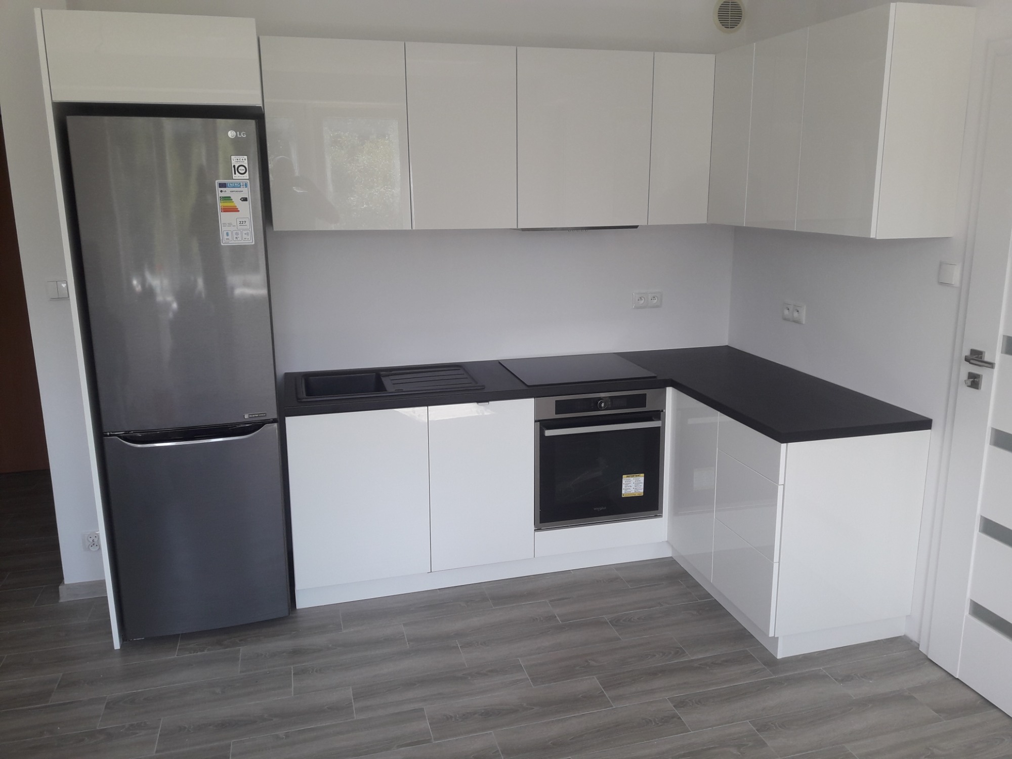 Nowoczesna, minimalistyczna kuchnia z białymi, lakierowanymi frontami, czarnym blatem i szarą lodówką LG, wykończona panelami podłogowymi imitującymi drewno.