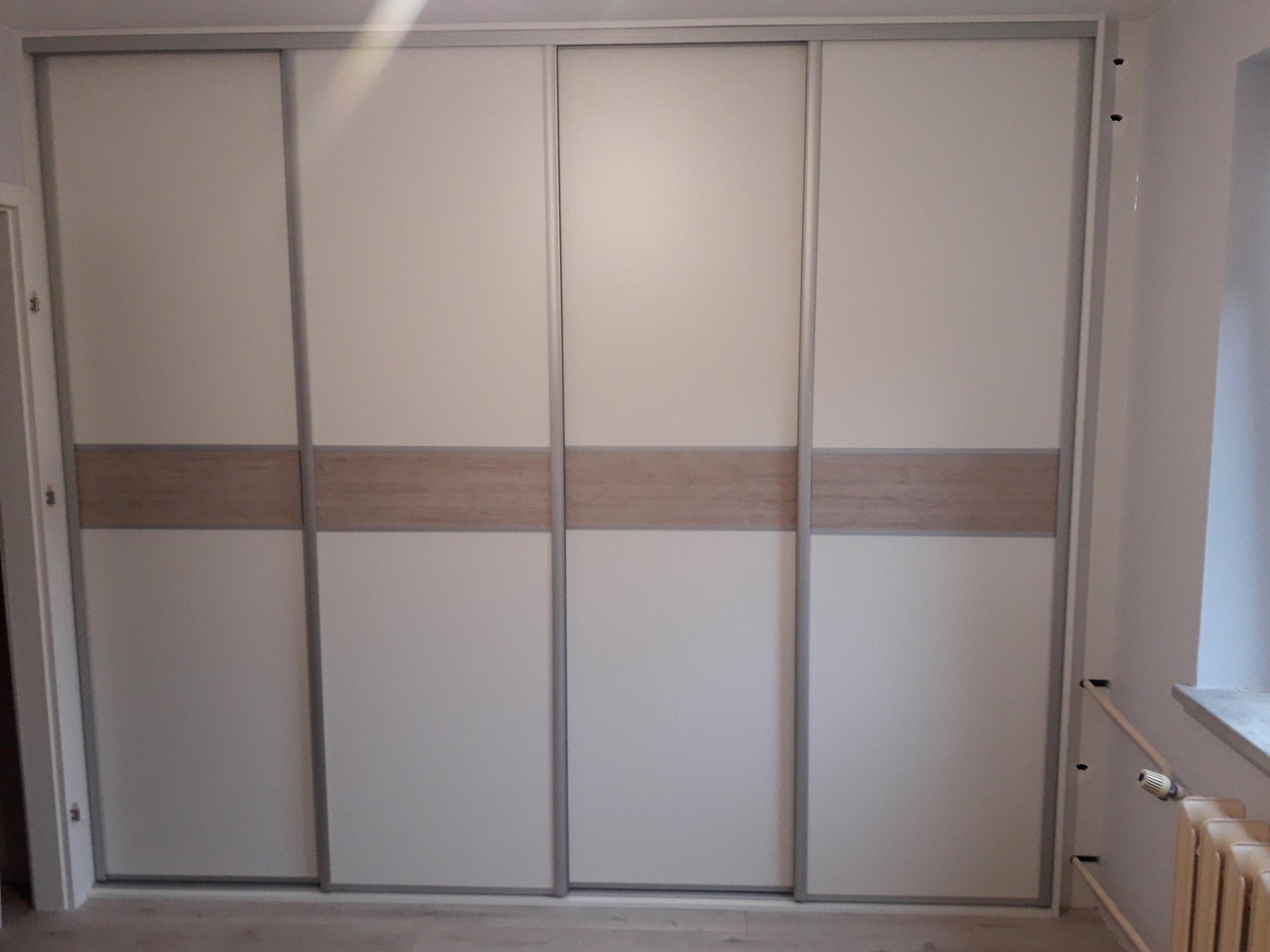 Czterodrzwiowa szafa przesuwna z białymi frontami i poziomym pasem w kolorze jasnego drewna, widoczny grzejnik obok.