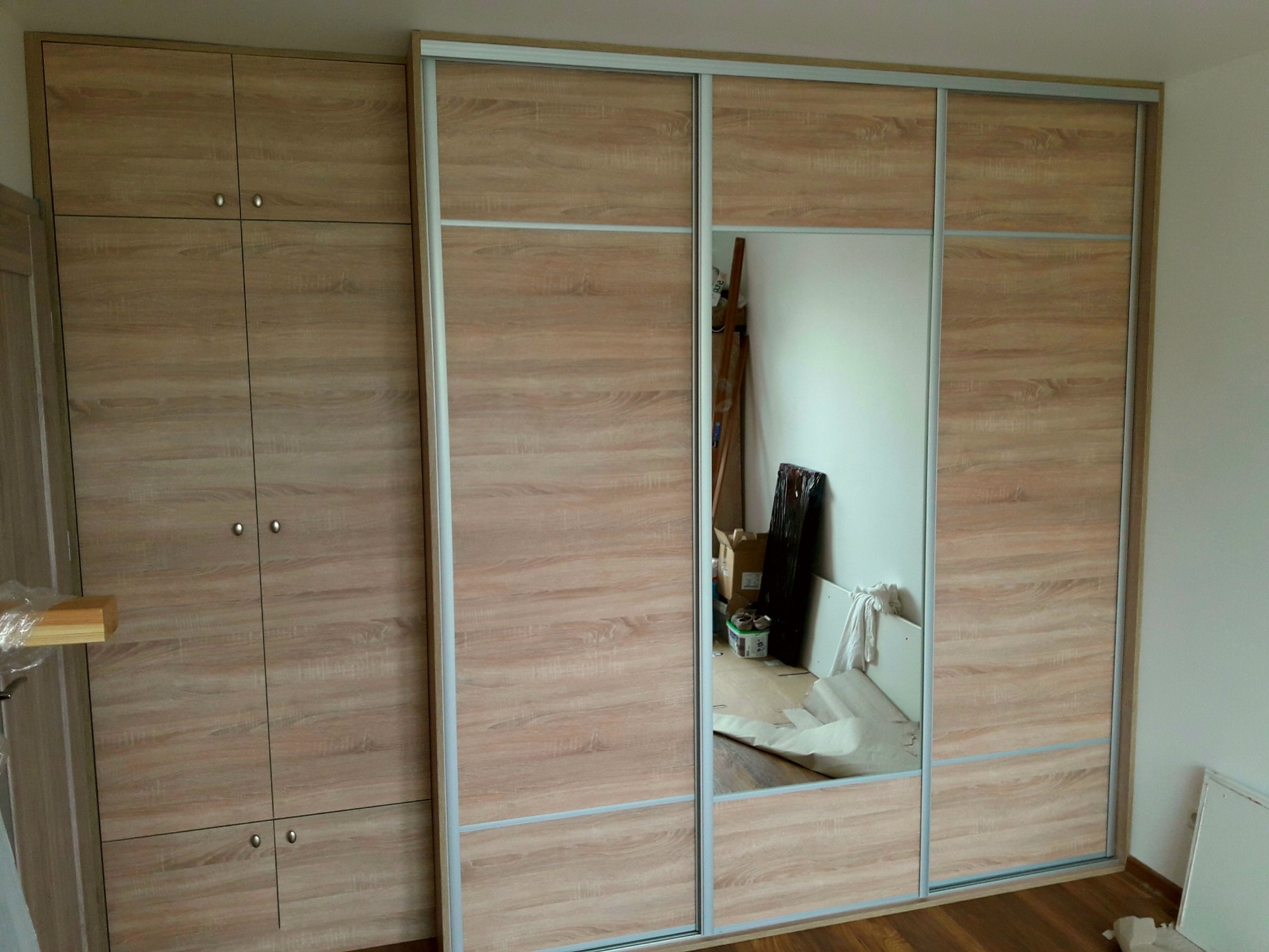 Duża szafa wnękowa z przesuwnymi drzwiami, wykończona okleiną imitującą jasne drewno, z lustrem w centralnej części, odbijającym fragment pokoju w trakcie prac wykończeniowych.
