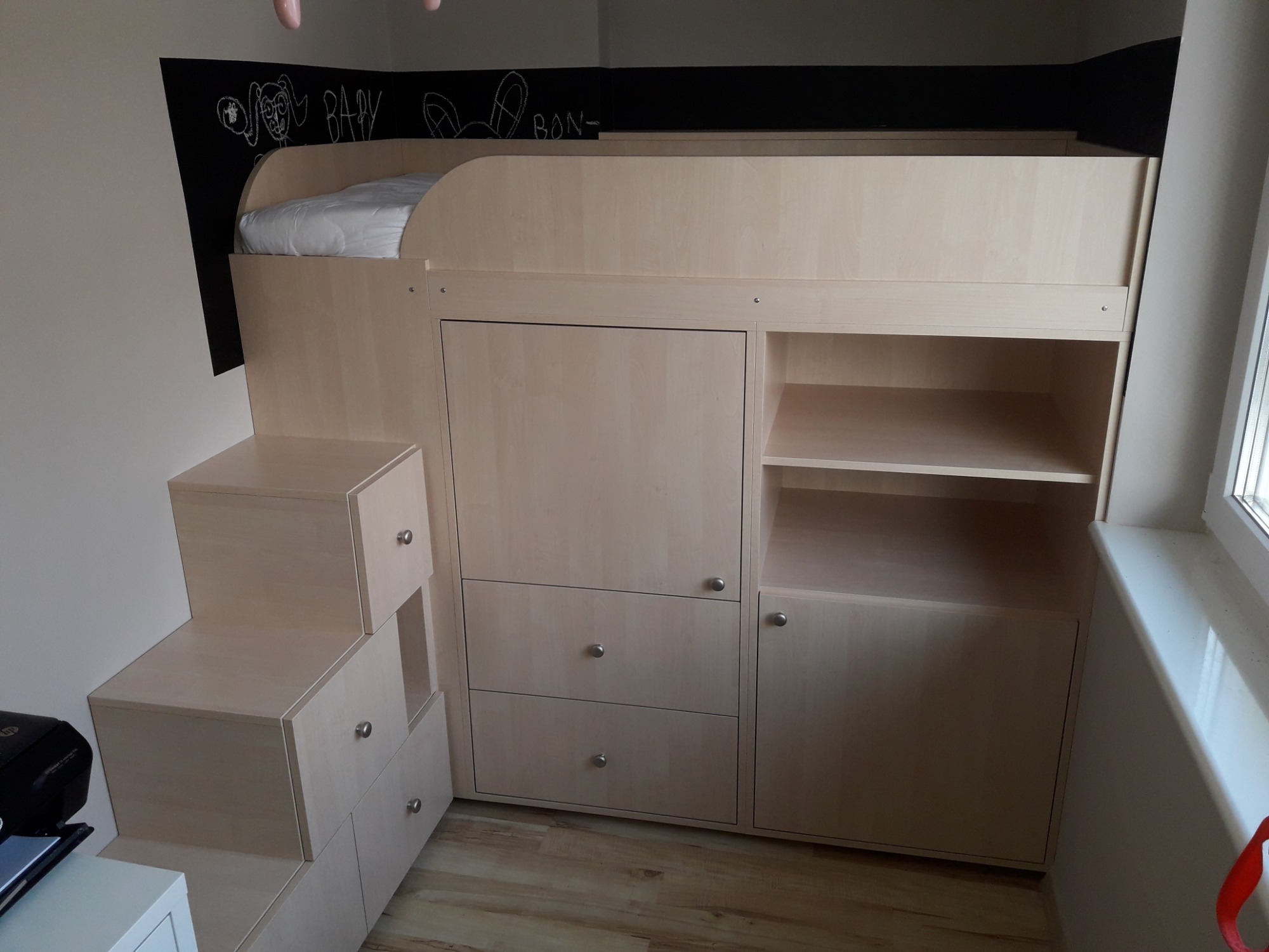 Białe łóżko piętrowe zintegrowane z szafkami i schodkami, rysunki kredą na czarnej ścianie w tle.