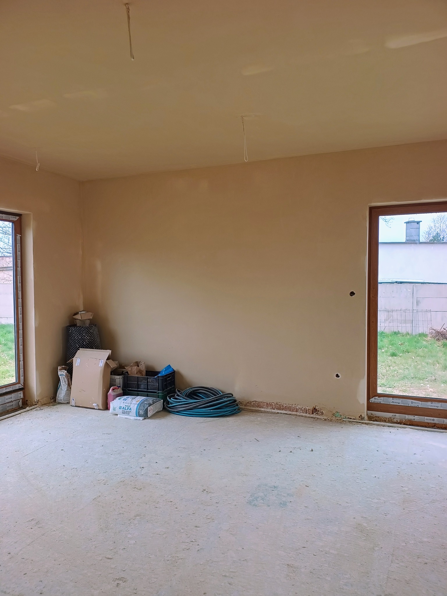 Wnętrze po tynkowaniu maszynowym: gładkie ściany w odcieniach beżu, widoczne worki z zaprawą i narzędzia, okna z widokiem na zieleń. Podłoga zakurzona.