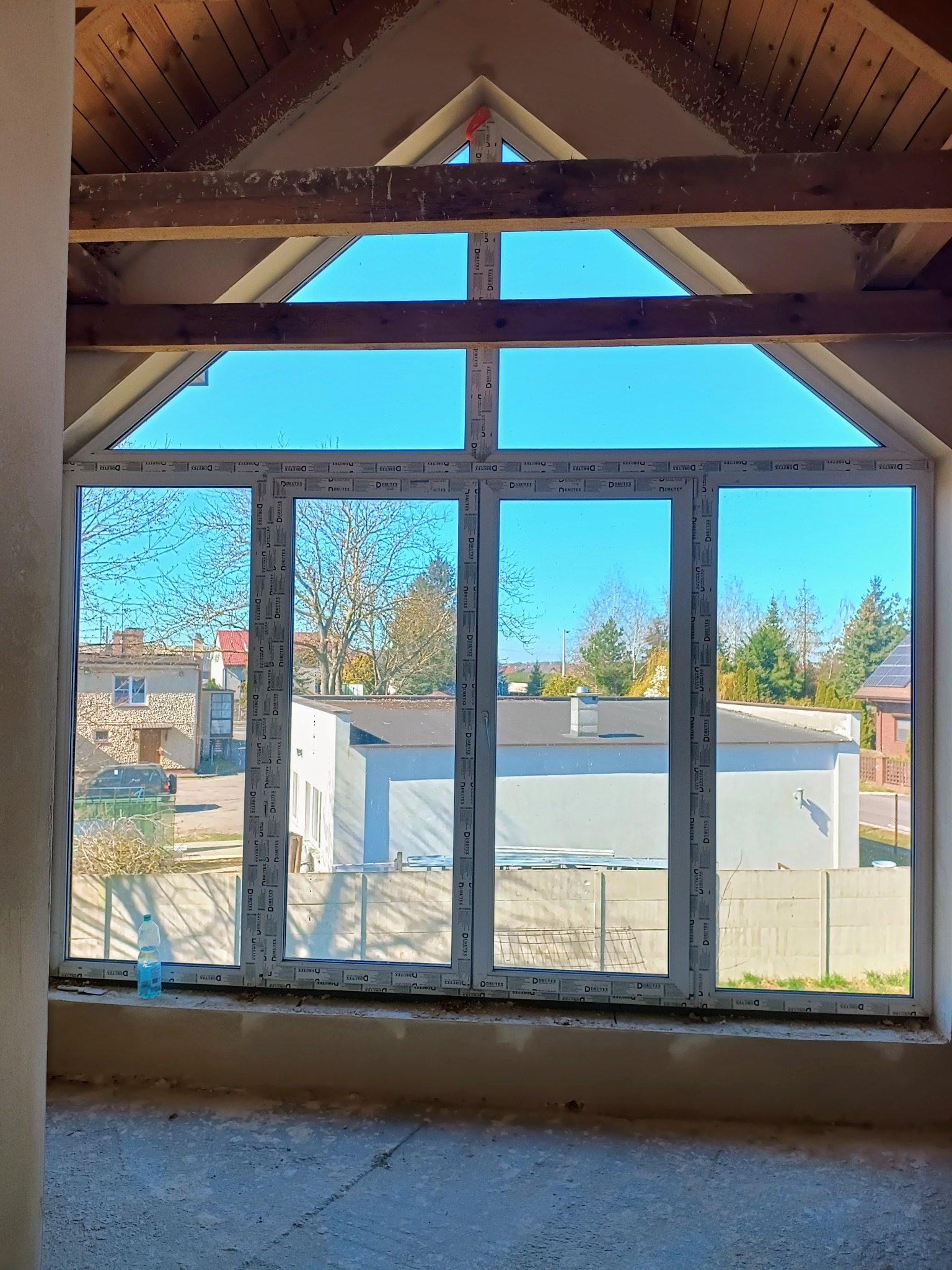 Nowe okno z szybami zespolonymi w surowym wnętrzu z drewnianą konstrukcją dachu. Widok na zabudowę jednorodzinną. Butelka wody na parapecie.