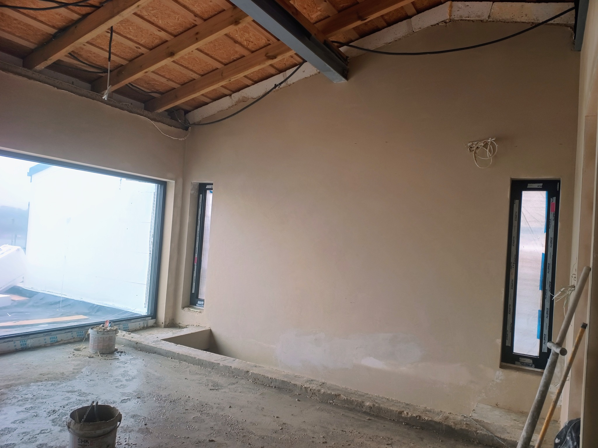 Wnętrze w trakcie prac tynkarskich: ściany pokryte świeżym tynkiem, widoczne okna z folią ochronną, surowe drewniane elementy konstrukcyjne dachu.
