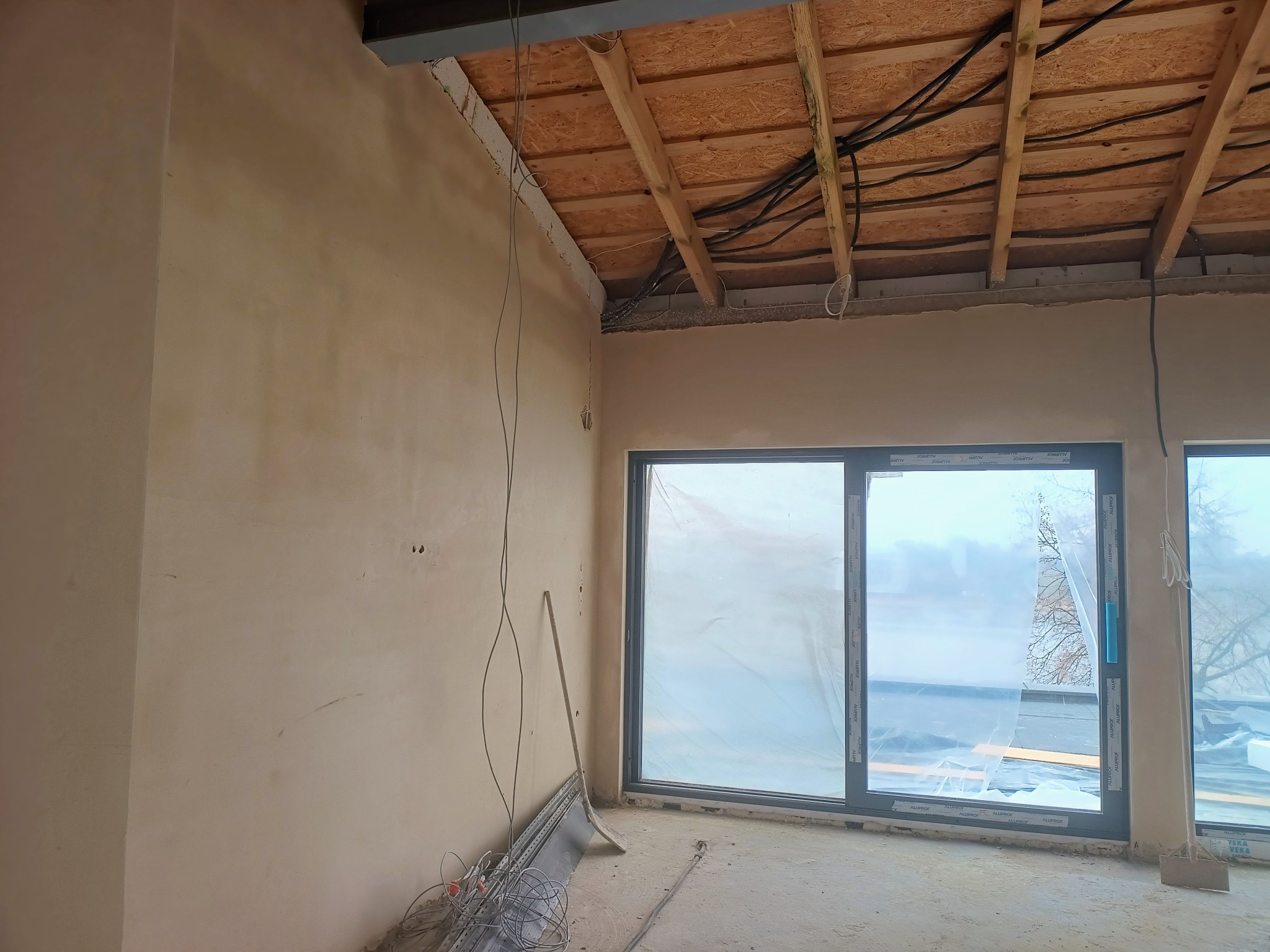 Ściana po tynkowaniu maszynowym z widocznym drewnianym stropem i oknami. Przewody elektryczne zwisają ze ściany. Na podłodze leżą narzędzia i materiały.
