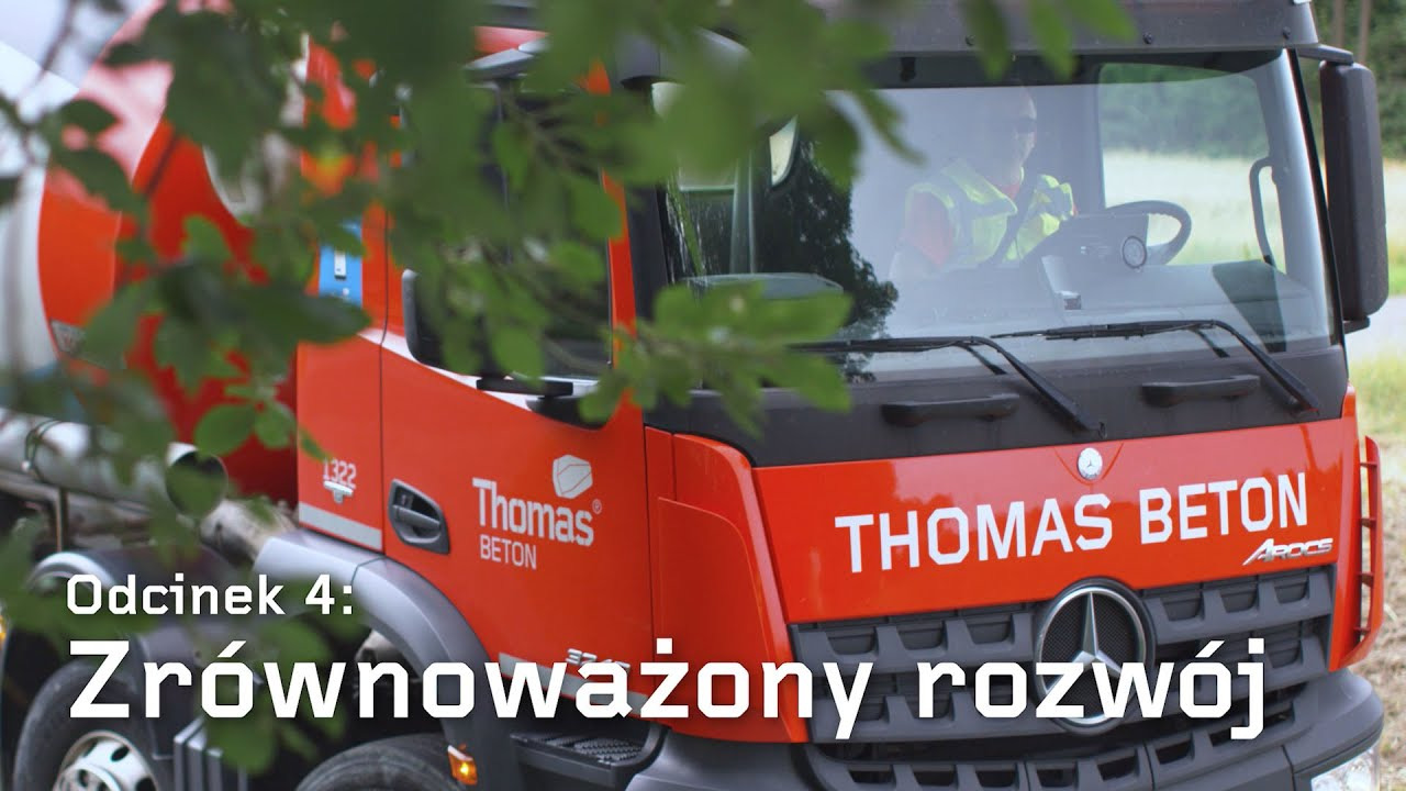 Czerwona betonomieszarka marki Thomas Beton z widocznym kierowcą w kamizelce odblaskowej, częściowo zasłonięta gałęziami drzewa.