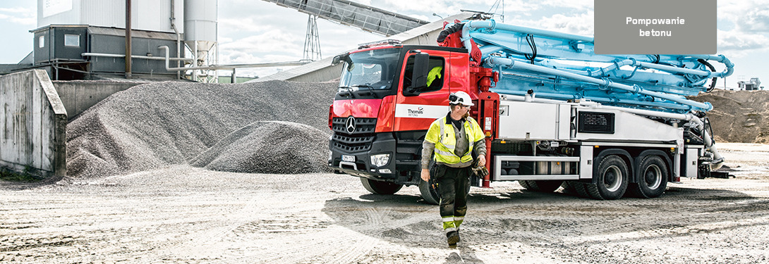 Ciężarówka do pompowania betonu z wysięgnikiem, pracownik w kasku i odblaskowej kamizelce idzie po placu budowy, w tle hałda kruszywa i zabudowania betoniarni.