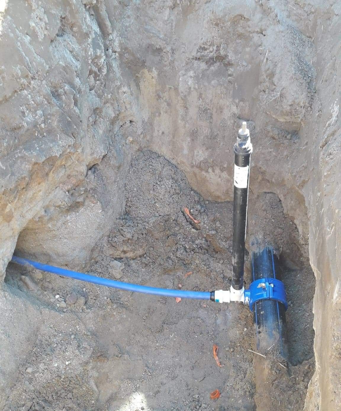 Niebieska rura wodna podłączona do czarnej rury w wykopie ziemnym z widocznymi złączkami.