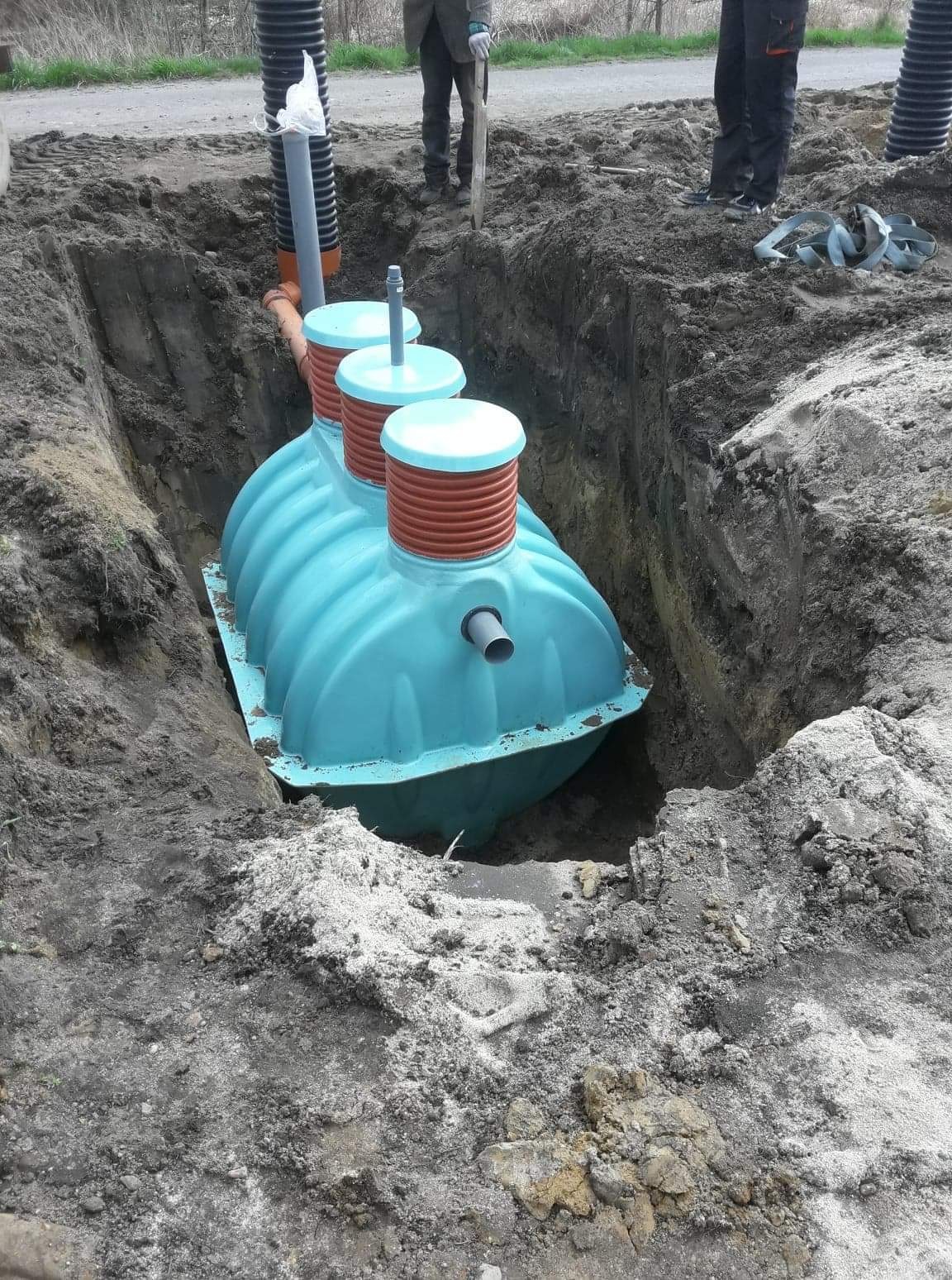 Niebieski zbiornik szamba betonowego z trzema włazami umieszczony w wykopie, widoczne rury i częściowo zasypana ziemia.
