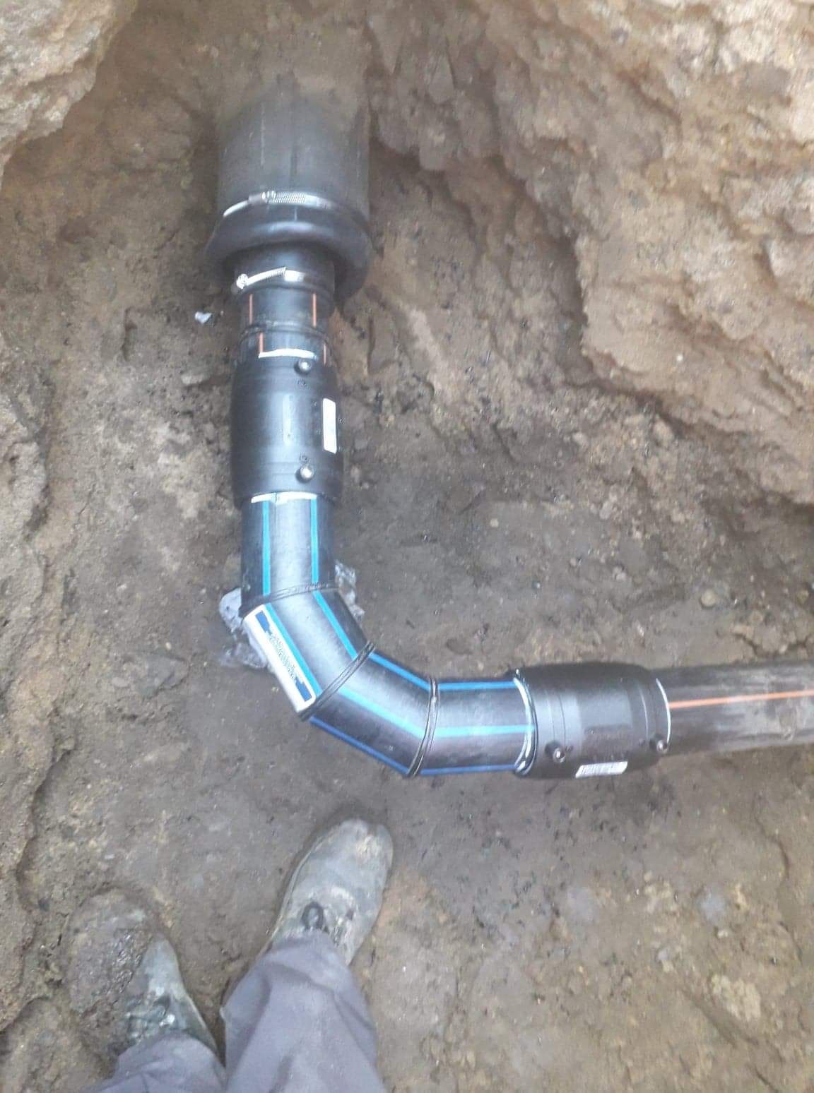 Czarny rurociąg z łącznikami, ułożony w wykopie ziemnym, widoczne buty robocze w dolnej części kadru.