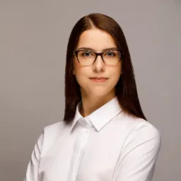 Portret młodej kobiety w okularach i białej koszuli na jednolitym, szarym tle. Profesjonalny wizerunek osoby, wzbudzający zaufanie.