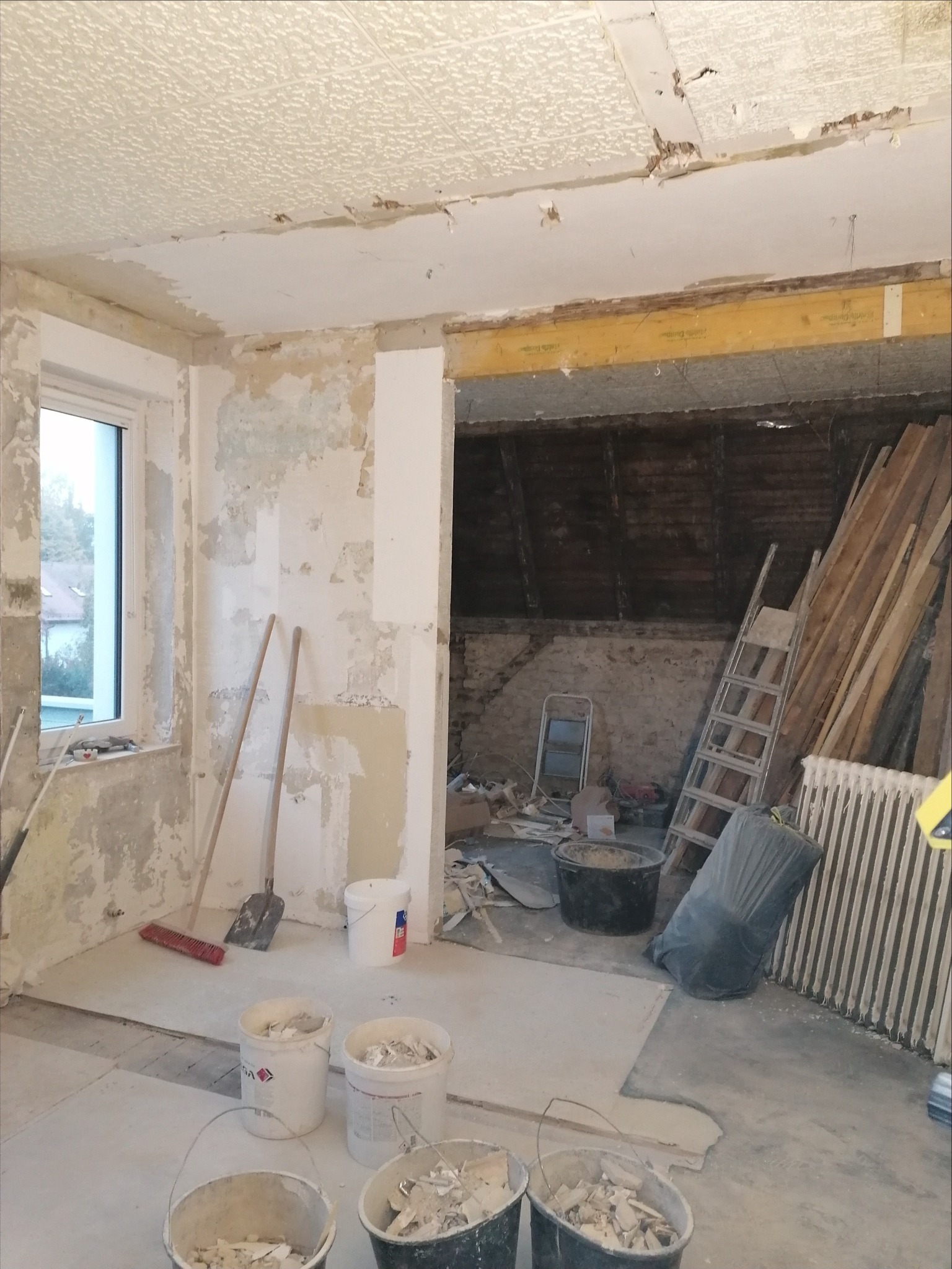 Wnętrze w trakcie demontażu starej warstwy wykończeniowej: ściany z resztkami tapet i farb, sufit z widocznymi uszkodzeniami, wiadra z gruzem, drabiny i narzędzia budowlane.