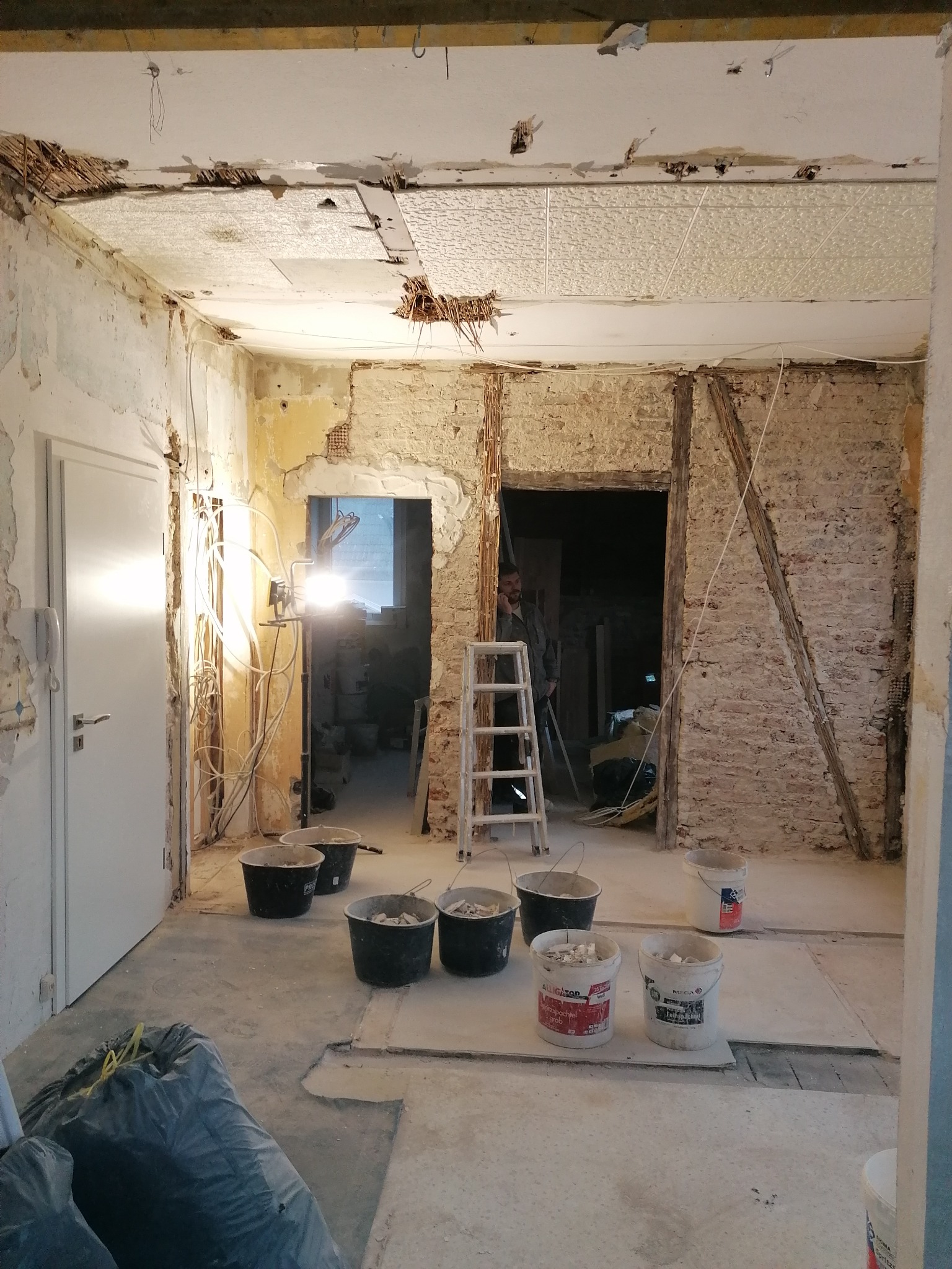 Demontaż starego sufitu i odsłonięta cegła na ścianach w trakcie remontu mieszkania, widoczne wiadro z gruzem, drabina i lampa budowlana