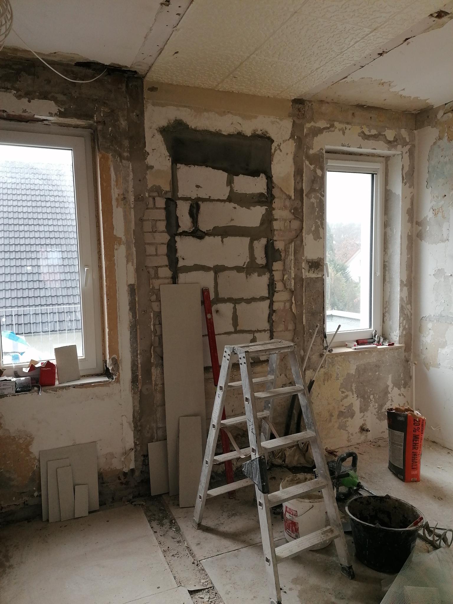 Pokój w trakcie prac remontowych: odsłonięta cegła, resztki starej farby na ścianach, drabina, narzędzia i materiały budowlane, okna z widokiem na zewnątrz.