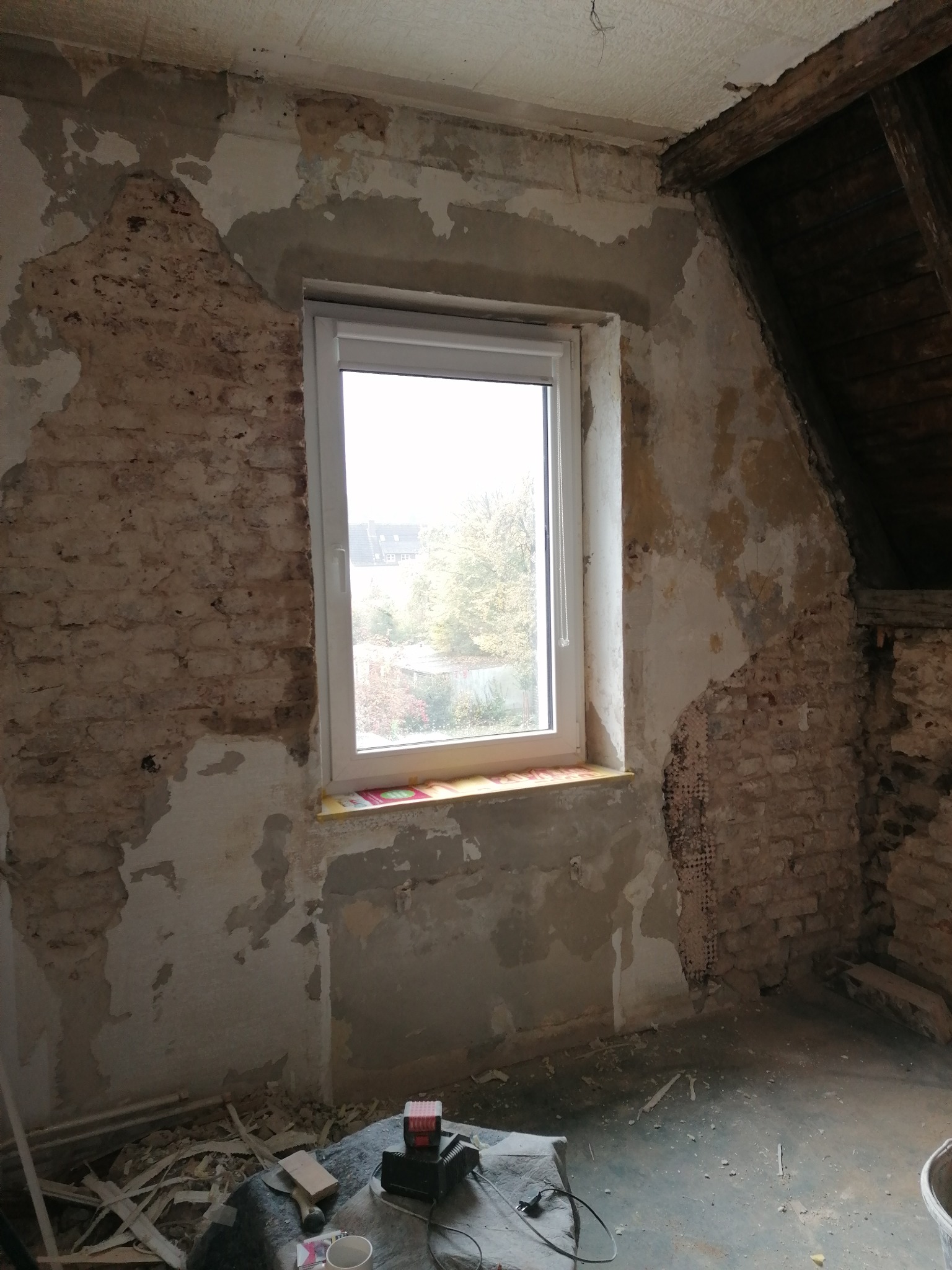 Remontowane poddasze w Kłodawie: odsłonięta cegła, fragment dachu z drewnianymi belkami, nowe okno PCV, ślady starej farby i gładzi na ścianach, narzędzia i resztki materiałów na podłodze.
