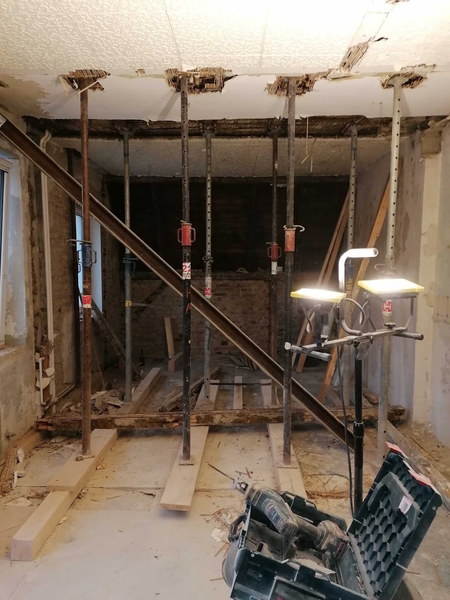 Wnętrze w trakcie demontażu sufitu, widoczne metalowe stemple podtrzymujące strop, odsłonięta konstrukcja, narzędzia w kuferku na podłodze, lampa budowlana.