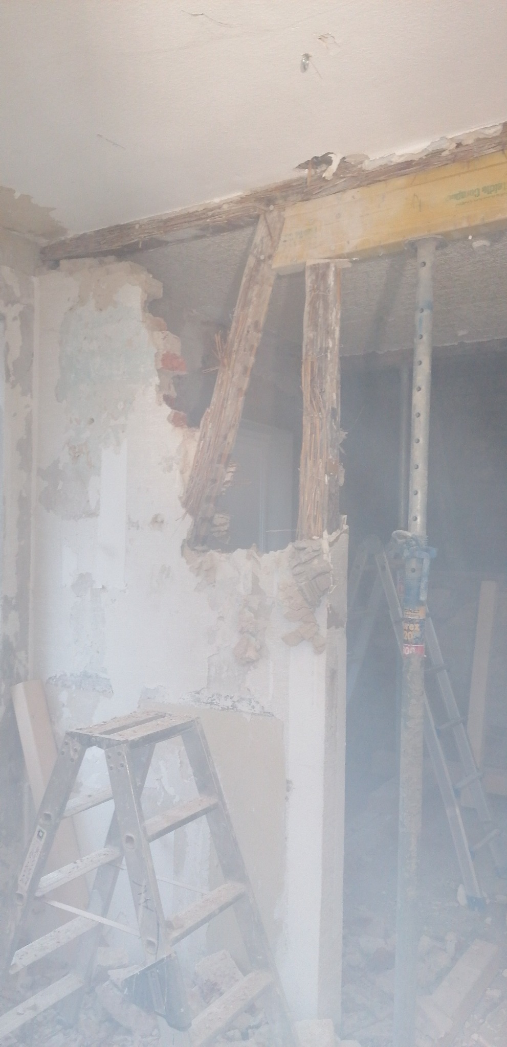 Fragment ściany z odsłoniętą konstrukcją drewnianą i ceglaną podczas prac remontowych, wspartą na metalowej podporze, z widoczną drabiną w zapylonym pomieszczeniu.