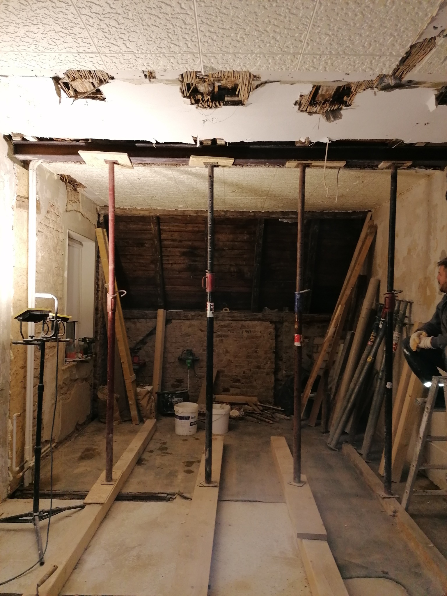 Wnętrze w trakcie przebudowy: odsłonięty strop z widocznymi elementami konstrukcyjnymi, podparty stalowymi słupami i drewnianymi belkami, z elementami instalacji elektrycznej i resztkami starego...