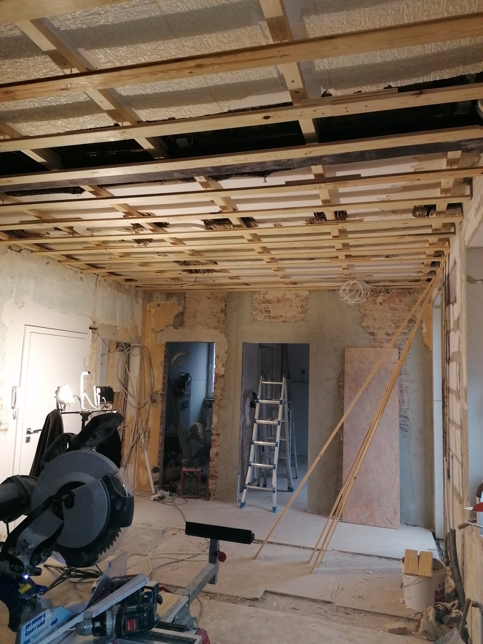 Drewniana konstrukcja sufitu podwieszanego w trakcie montażu, widoczne ocieplenie, przewody elektryczne, piła ukośnica i drabina w pomieszczeniu w trakcie prac.