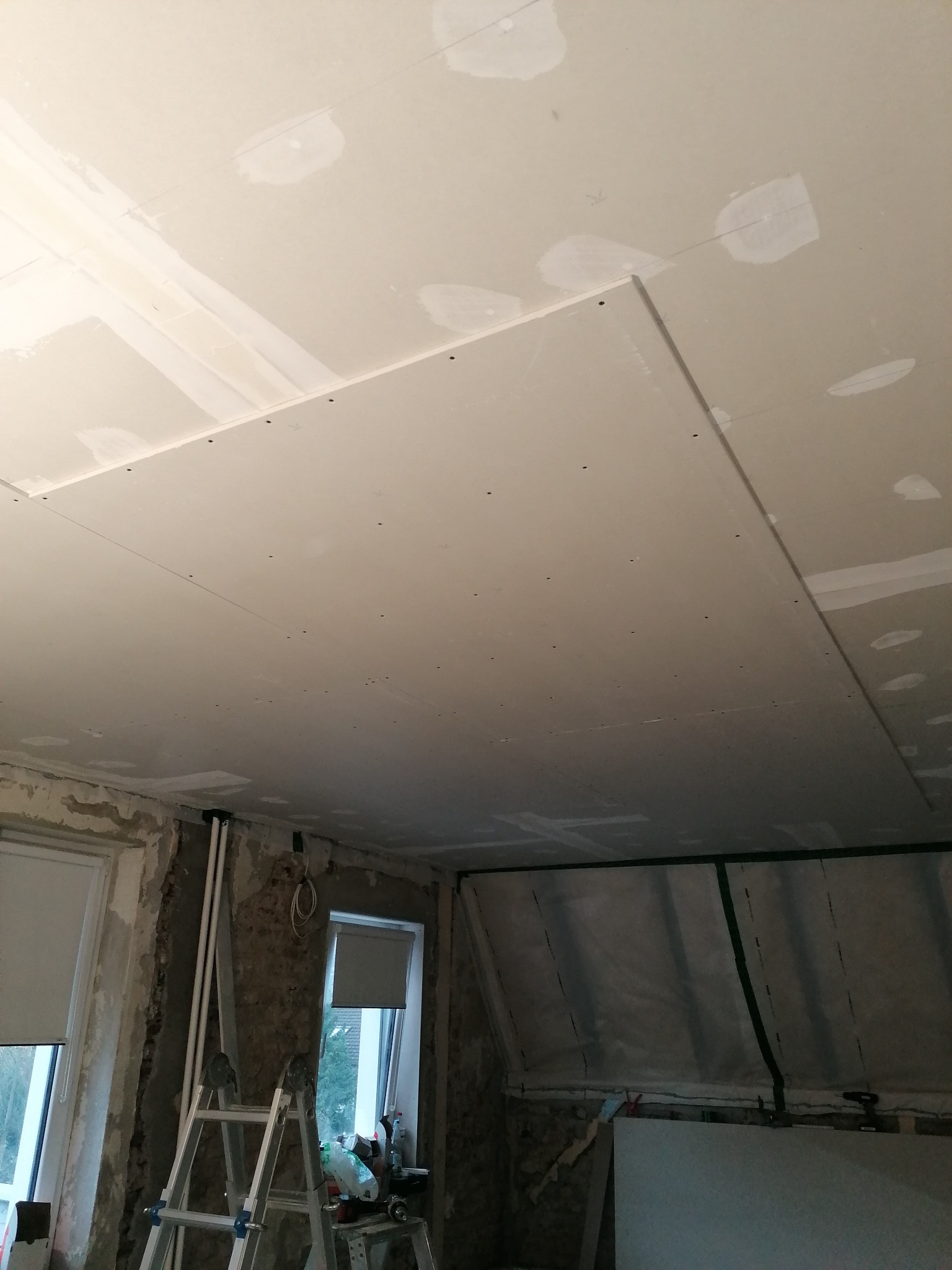 Montaż płyt gipsowo-kartonowych na suficie, widoczne łączenia płyt, ślady szpachlowania, drabina i okna w tle.