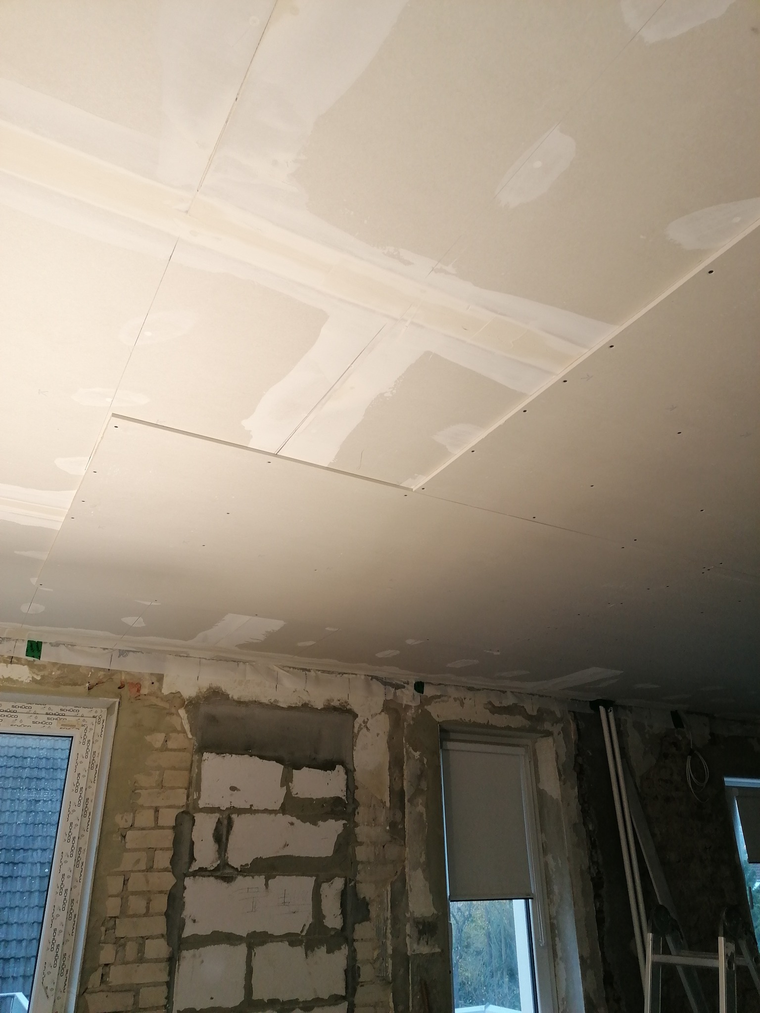 Montaż płyt gipsowo-kartonowych na suficie, widoczne szpachlowane łączenia i niedokończone fragmenty sufitu, ściany w trakcie przygotowania pod wykończenie, okna PCV.