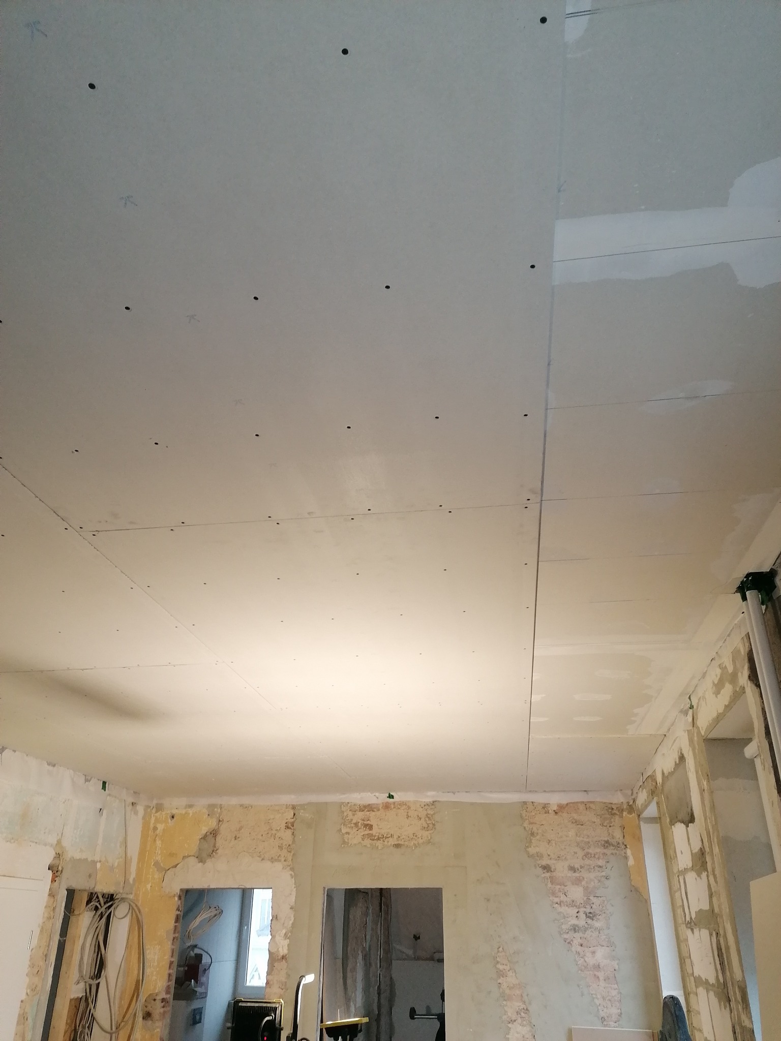 Montaż płyt gipsowo-kartonowych na suficie w trakcie prac wykończeniowych, widoczne ślady po wierceniu i ołówkowe oznaczenia na płytach, surowe ściany w tle.
