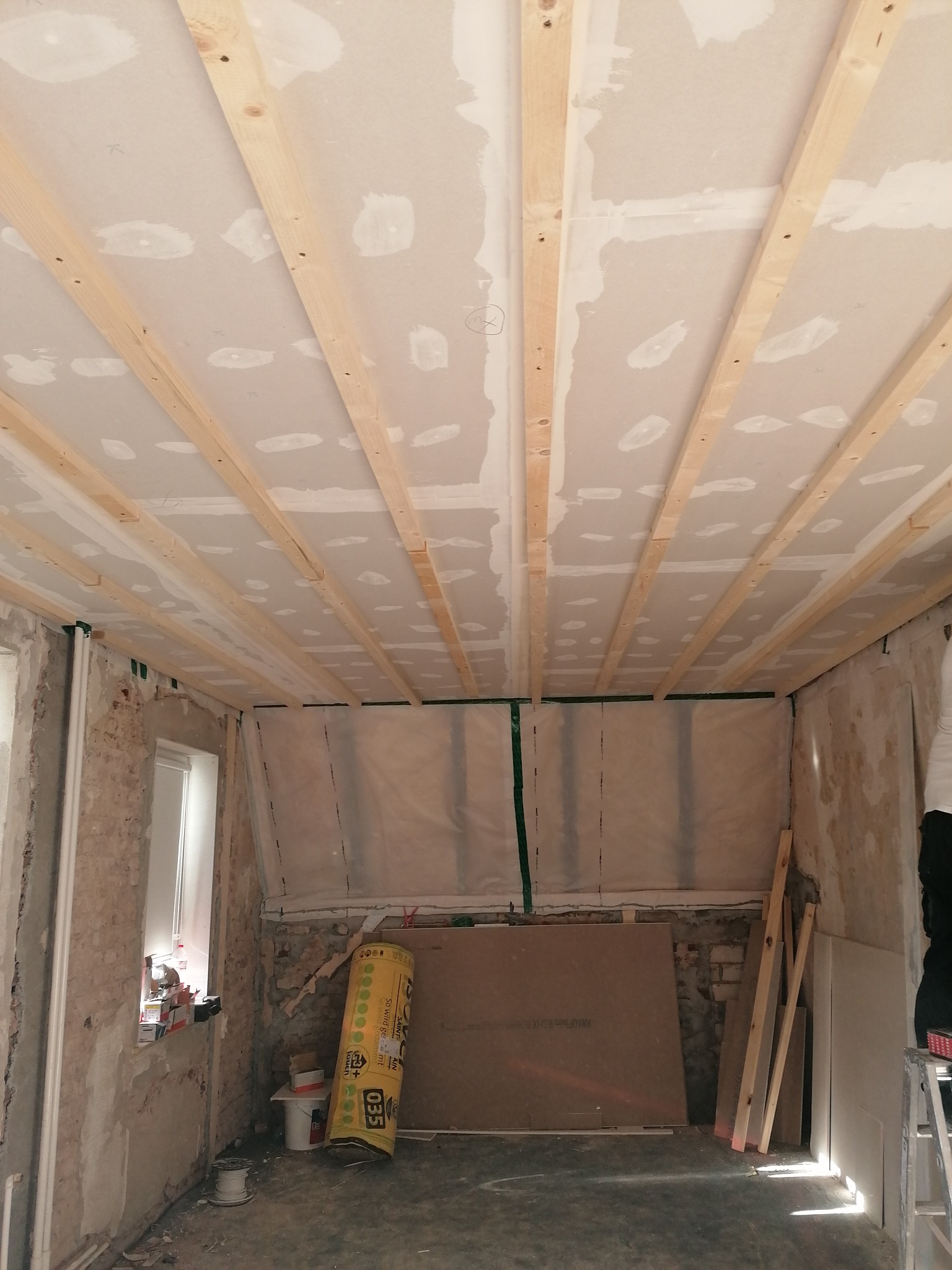 Wnętrze w trakcie prac: drewniana konstrukcja sufitu podwieszanego, częściowo pokryta płytami gipsowo-kartonowymi z widocznymi łączeniami, surowe ściany z fragmentami starego muru, materiały...