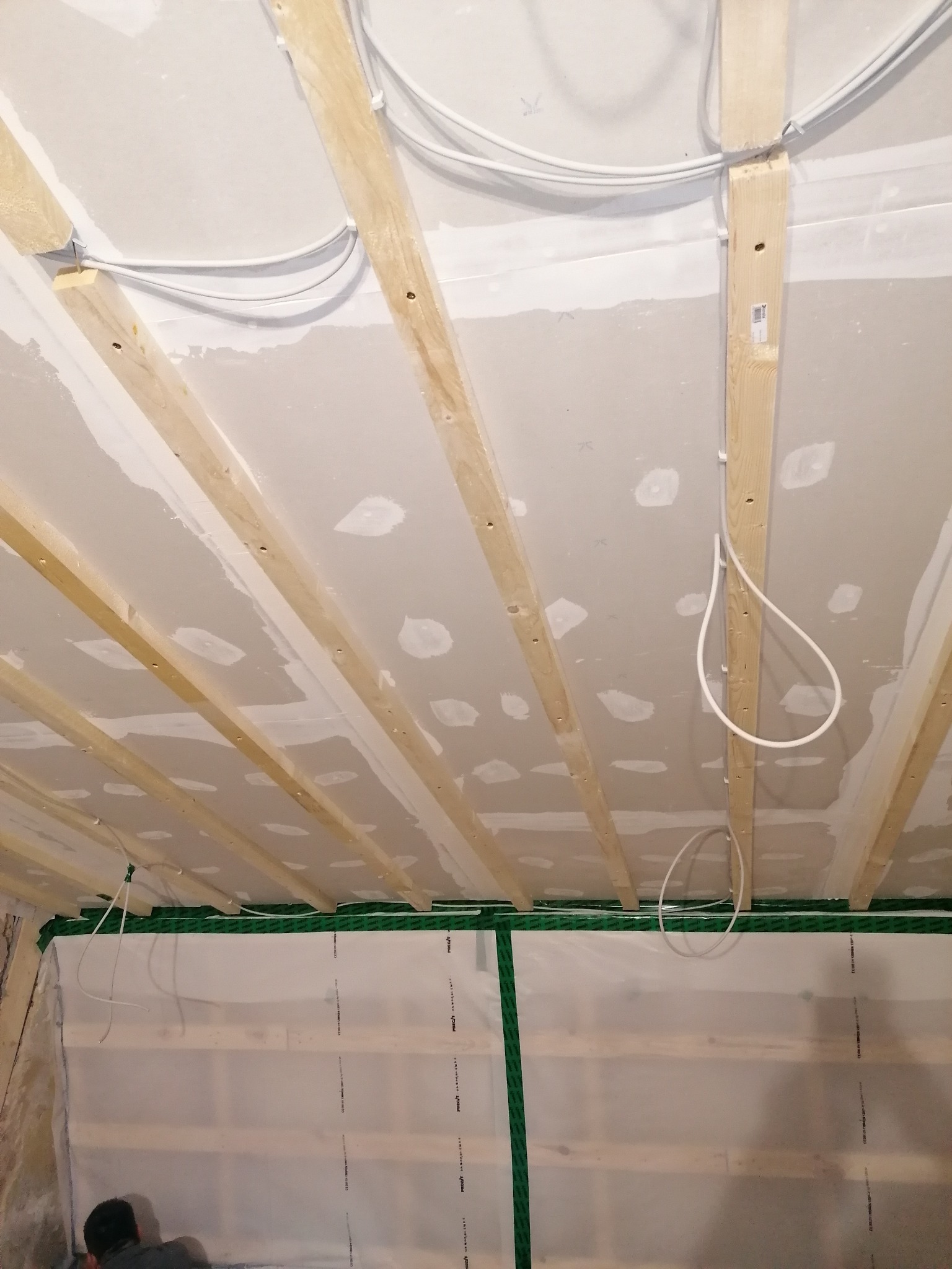 Sufit w trakcie prac wykończeniowych, widoczne drewniane belki konstrukcyjne, instalacja elektryczna oraz płyty gipsowo-kartonowe z miejscowymi szpachlowaniami, zabezpieczenie folią malarską dolnej...