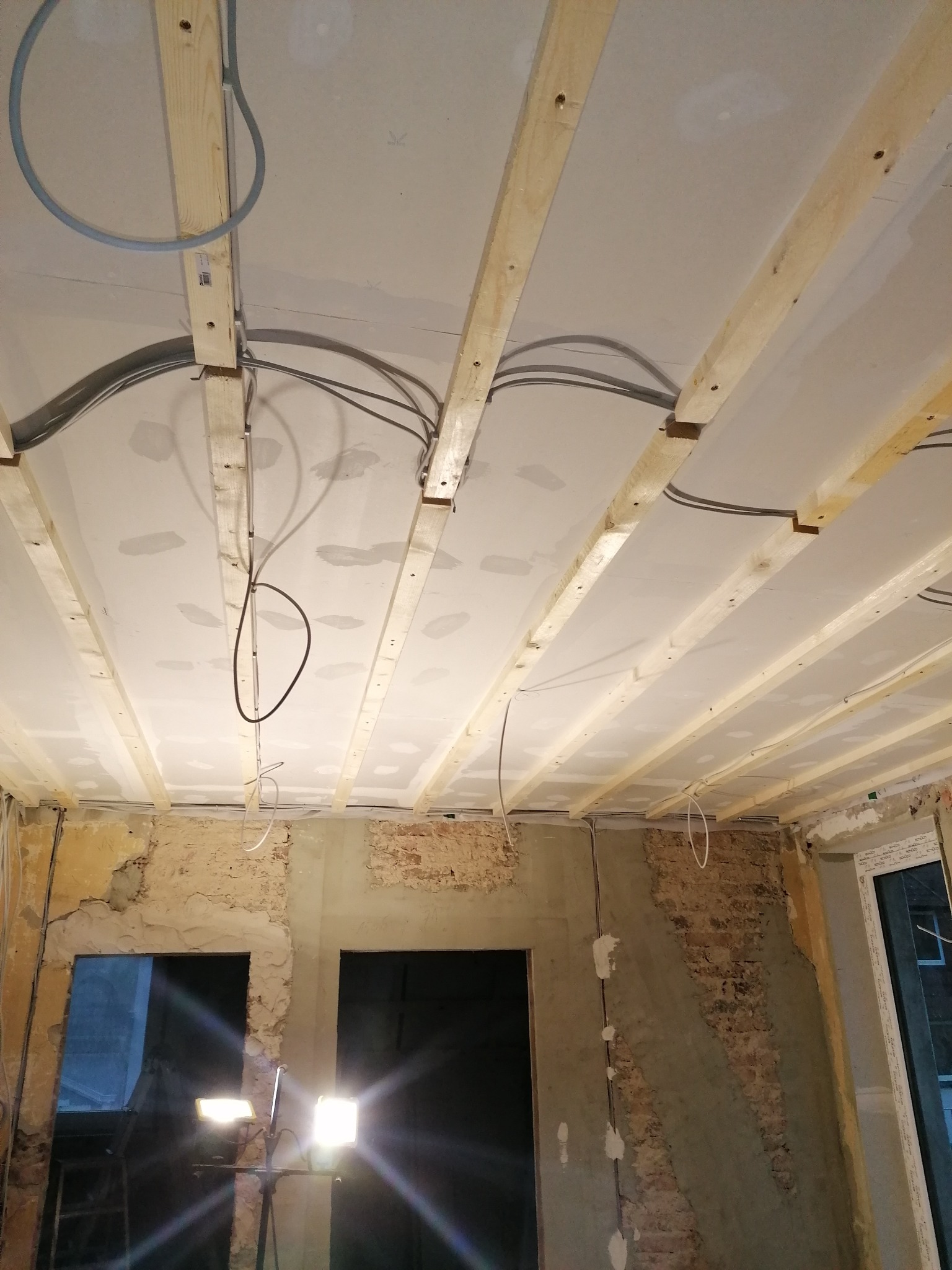 Wnętrze w trakcie prac przygotowawczych do wykończenia: surowy sufit z drewnianą konstrukcją, odsłonięte fragmenty cegieł na ścianach, widoczne instalacje elektryczne.