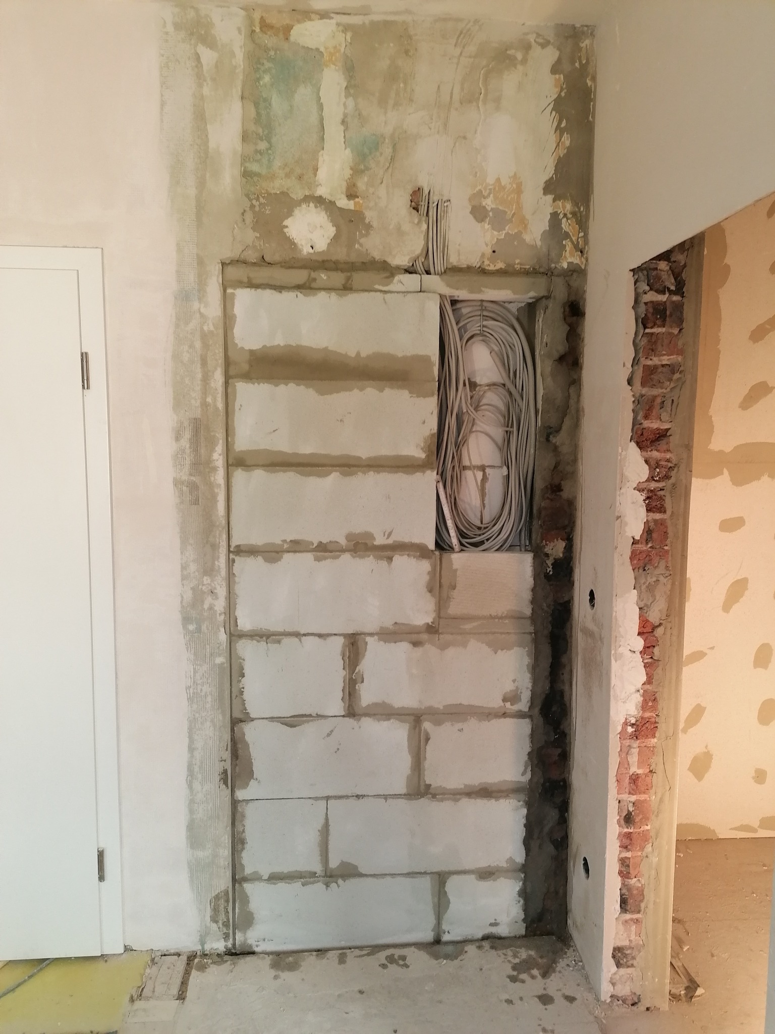 Wnęka w ścianie z bloków betonowych, widoczne przewody elektryczne, surowy stan wykończenia, fragment ściany z odsłoniętą czerwoną cegłą.