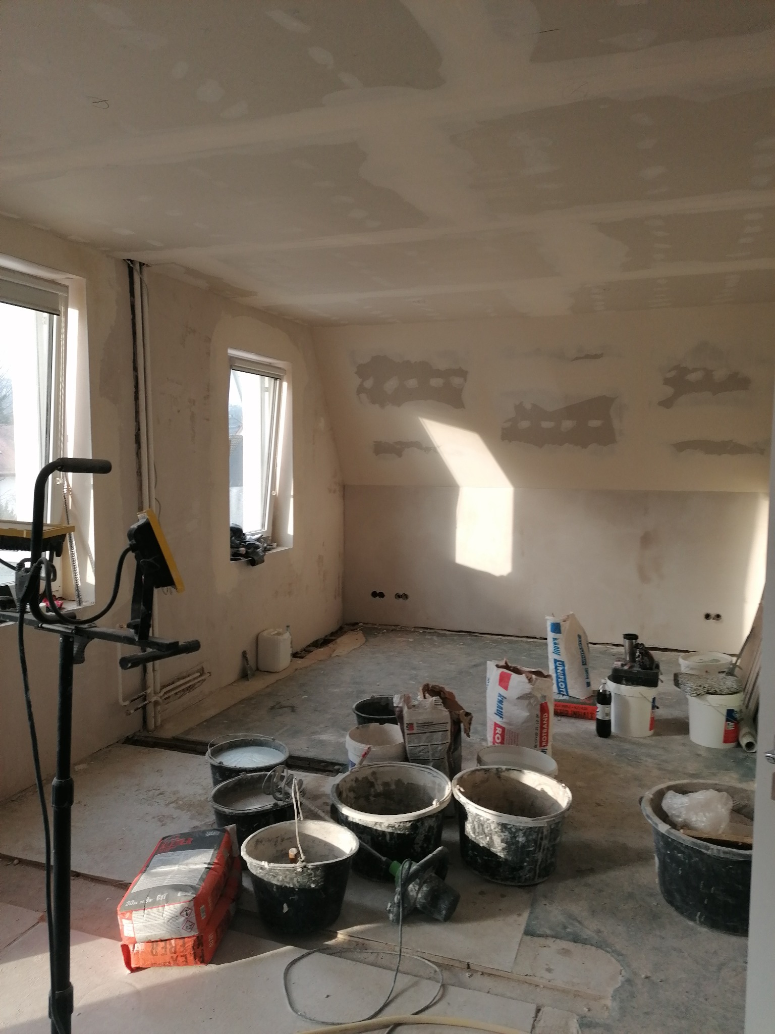 Pokój w trakcie prac wykończeniowych: ściany częściowo pokryte gładzią, widoczne worki z zaprawą, wiadra z resztkami materiałów, lampa budowlana na statywie.