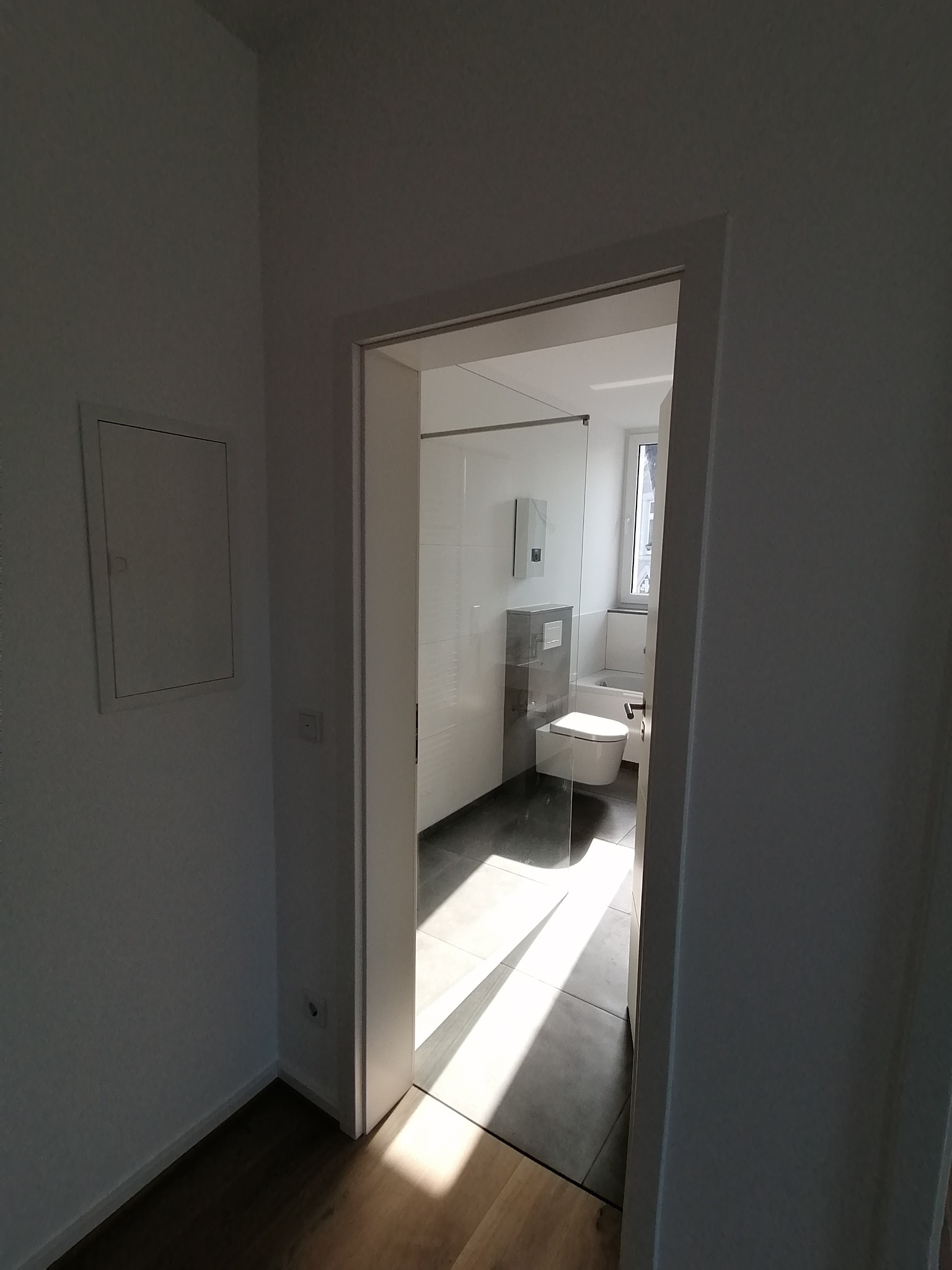 Widok przez drzwi na minimalistyczną łazienkę z kabiną prysznicową, toaletą i oknem, oświetloną promieniami słonecznymi padającymi na szare płytki podłogowe.