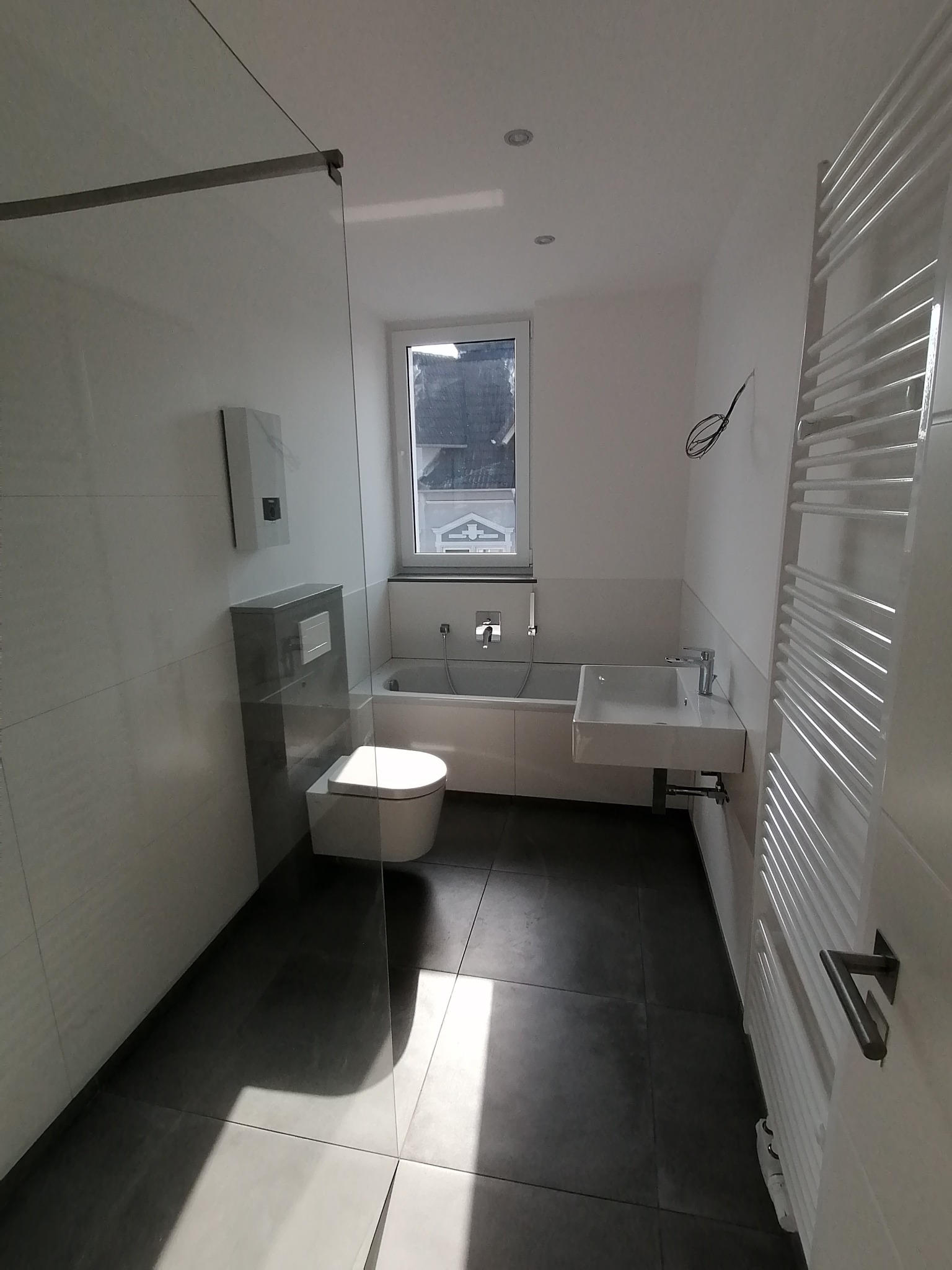 Nowoczesna łazienka z szarą podłogą, przeszkloną kabiną prysznicową, podwieszaną toaletą, białą wanną i grzejnikiem drabinkowym; widoczne okno z widokiem na budynki.