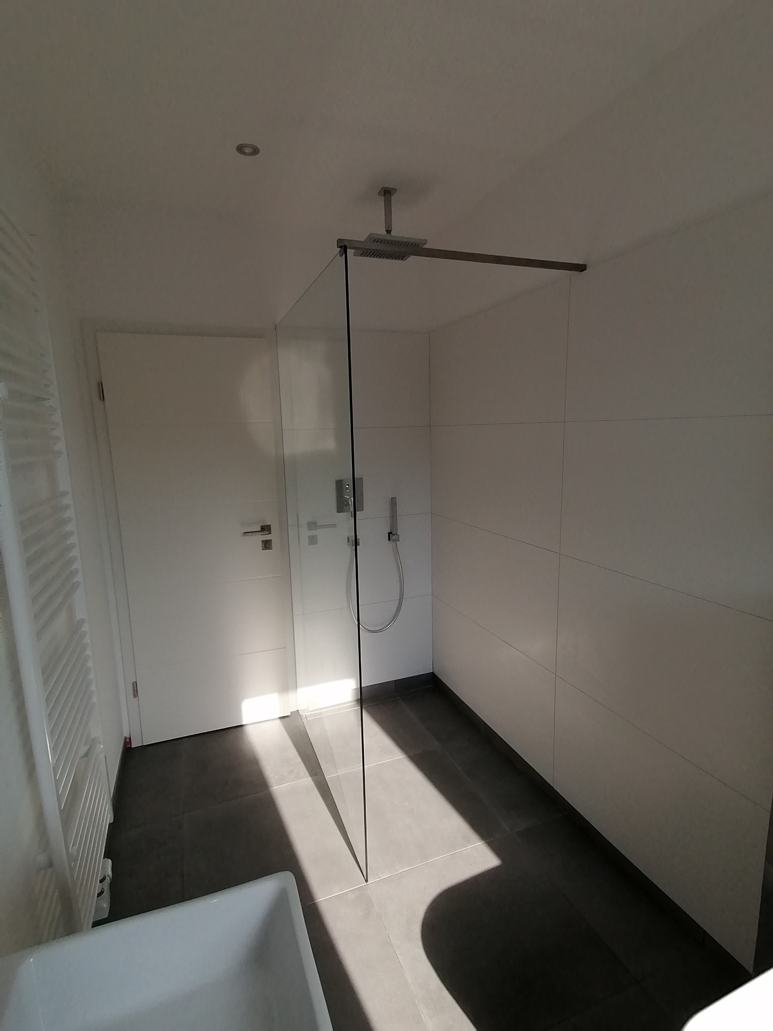 Minimalistyczna łazienka z kabiną prysznicową typu walk-in, szarymi płytkami podłogowymi i białymi płytkami ściennymi, widoczny grzejnik drabinkowy i fragment wanny.