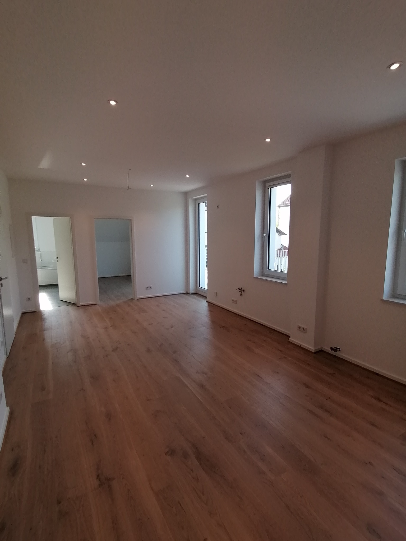 Jasne, przestronne wnętrze po remoncie z drewnianą podłogą, białymi ścianami, oknami i punktowym oświetleniem sufitowym, widok na sąsiednie pomieszczenia.