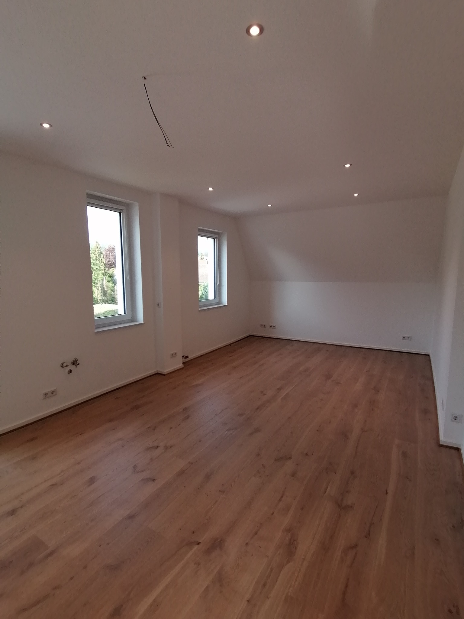 Puste, jasne pomieszczenie z drewnianą podłogą, białymi ścianami i oknami, widoczne wystające przewody z sufitu przed instalacją lampy.