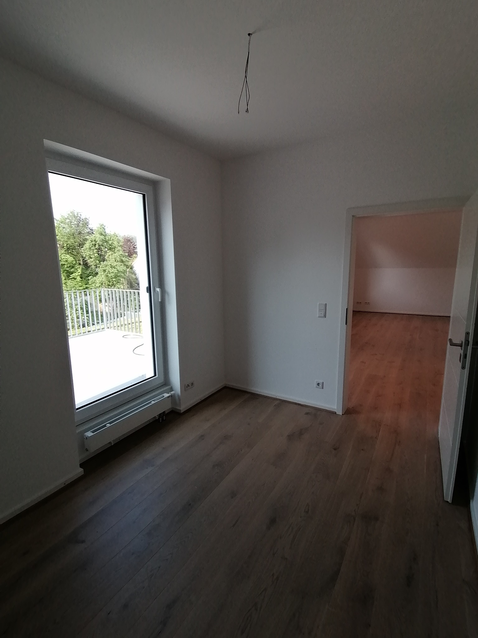 Pusty pokój z drewnianą podłogą, widok na balkon przez okno, surowe ściany przygotowane do malowania, widoczne kable wystające z sufitu.