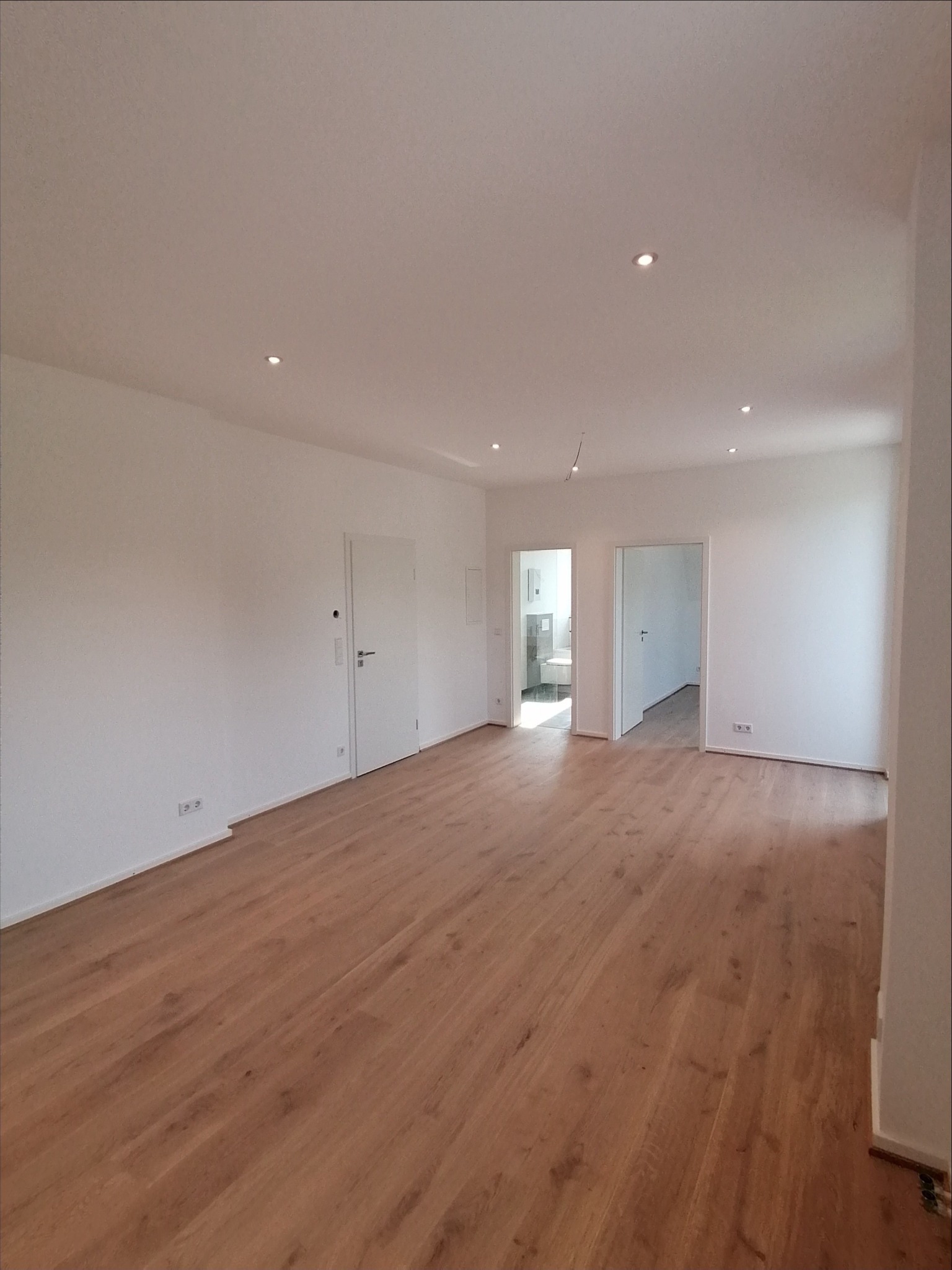 Jasne, przestronne wnętrze po remoncie, z drewnianą podłogą, białymi ścianami i sufitami z wbudowanymi punktowymi lampami, widoczne drzwi wewnętrzne i wejście do łazienki.