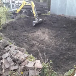 Żółta mini koparka wyrównuje teren pod przyszły staw, obok sterta cegieł i zaparkowana ciężarówka.