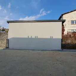 Świeżo pomalowana, gładka elewacja budynku w odcieniach szarości z widocznymi cieniami wystających element&oacute;w, sąsiadująca ze starszym budynkiem z widocznymi śladami napraw i roślinnością.