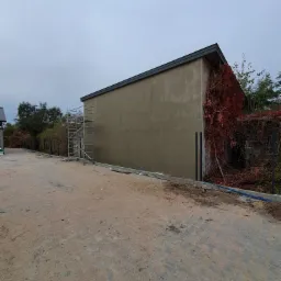 Ściana budynku po nałożeniu gładzi w Brańszczyku, widoczne rusztowanie i pnącza na sąsiedniej ścianie.