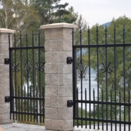 Czarny metalowy płot z ozdobnymi elementami i kamiennymi słupkami na tle zieleni i jeziora.