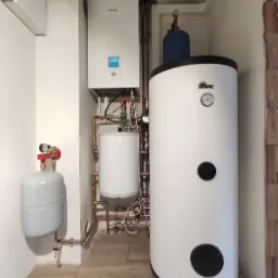 Zamontowany system grzewczy z pompą ciepła Panasonic i zasobnikiem Kospel, widoczne liczne miedziane rury i armatura, częściowo wykończone pomieszczenie.