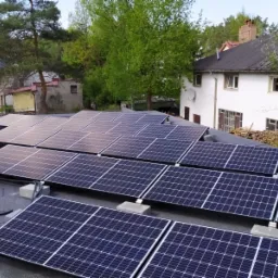 Instalacja paneli słonecznych na płaskim dachu budynku mieszkalnego, widoczne kable i narzędzia, w tle dom z białą elewacją i drzewa.