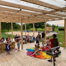 Animator w cylindrze i czerwonej koszuli tworzy balonowe zwierzątka dla dzieci siedzących na tęczowej płachcie, w tle rodzice na paletowych sofach pod drewnianą pergolą, widok na zielone pole i basen.