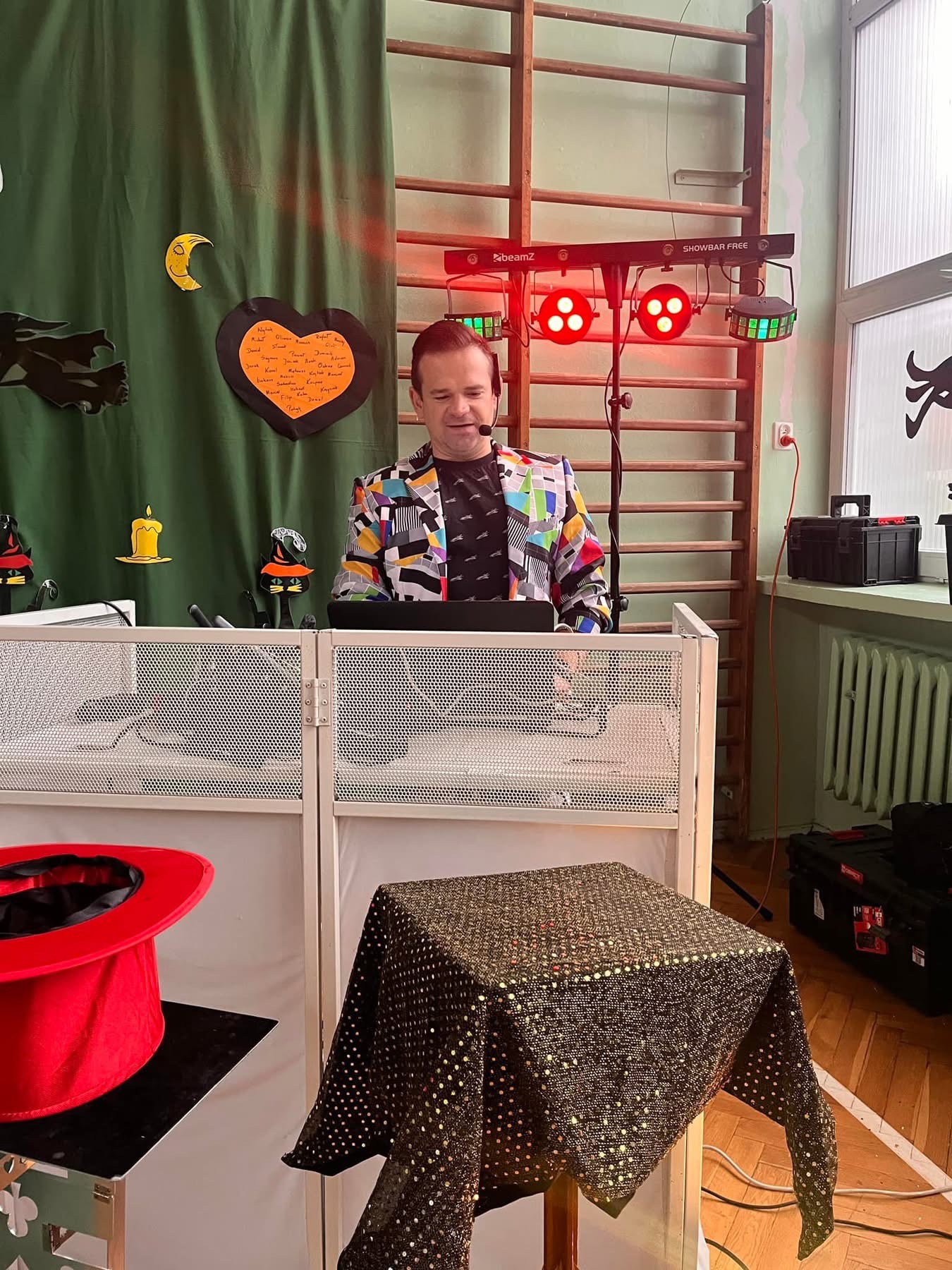 Mężczyzna w kolorowej marynarce z mikrofonem na głowie, za białym parawanem, z rekwizytami iluzjonistycznymi, w tle drabinka gimnastyczna i dekoracje halloweenowe na zielonym tle.